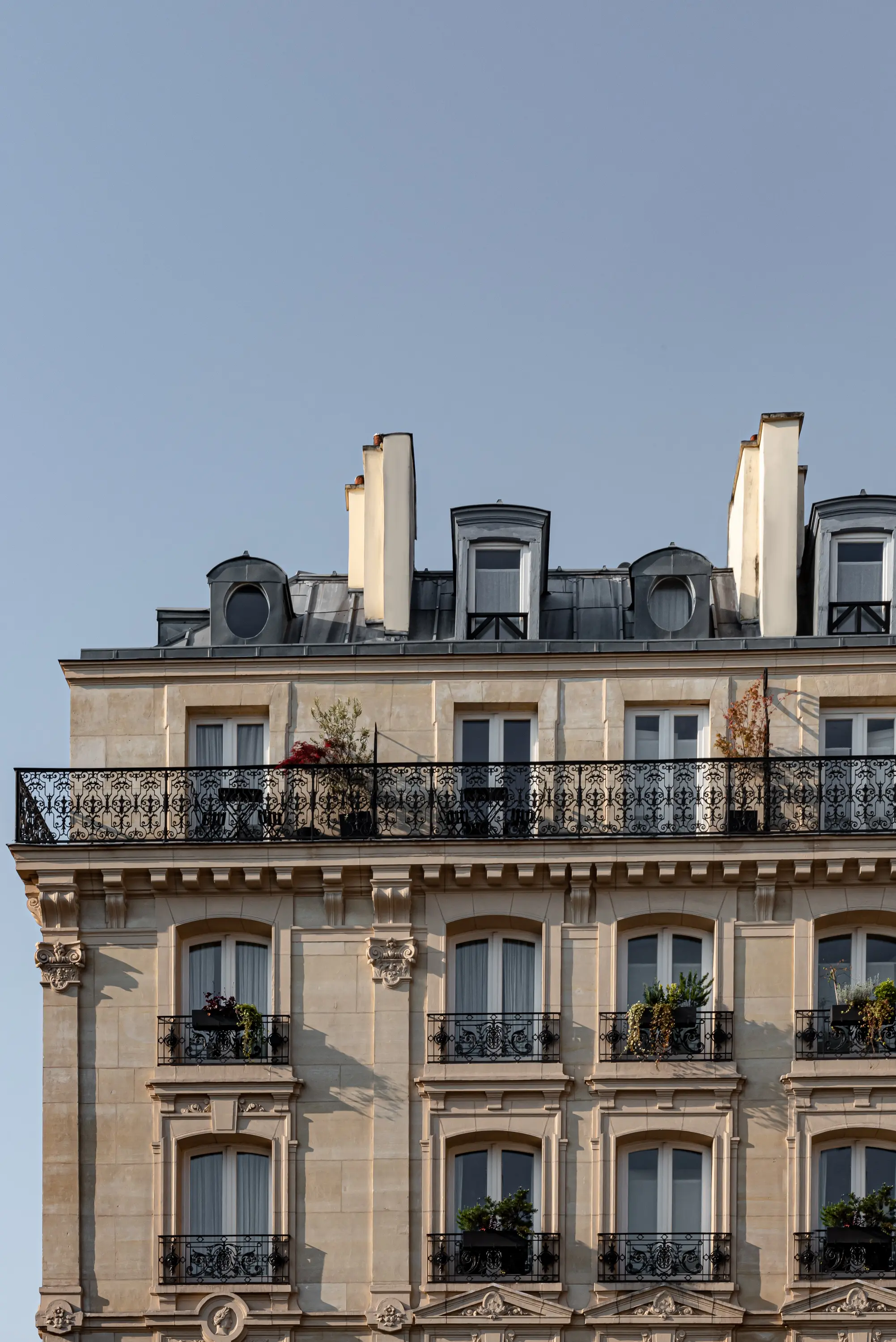 Classic beige stone building with black wrought-iron balconies and potted plants against a blue sky.