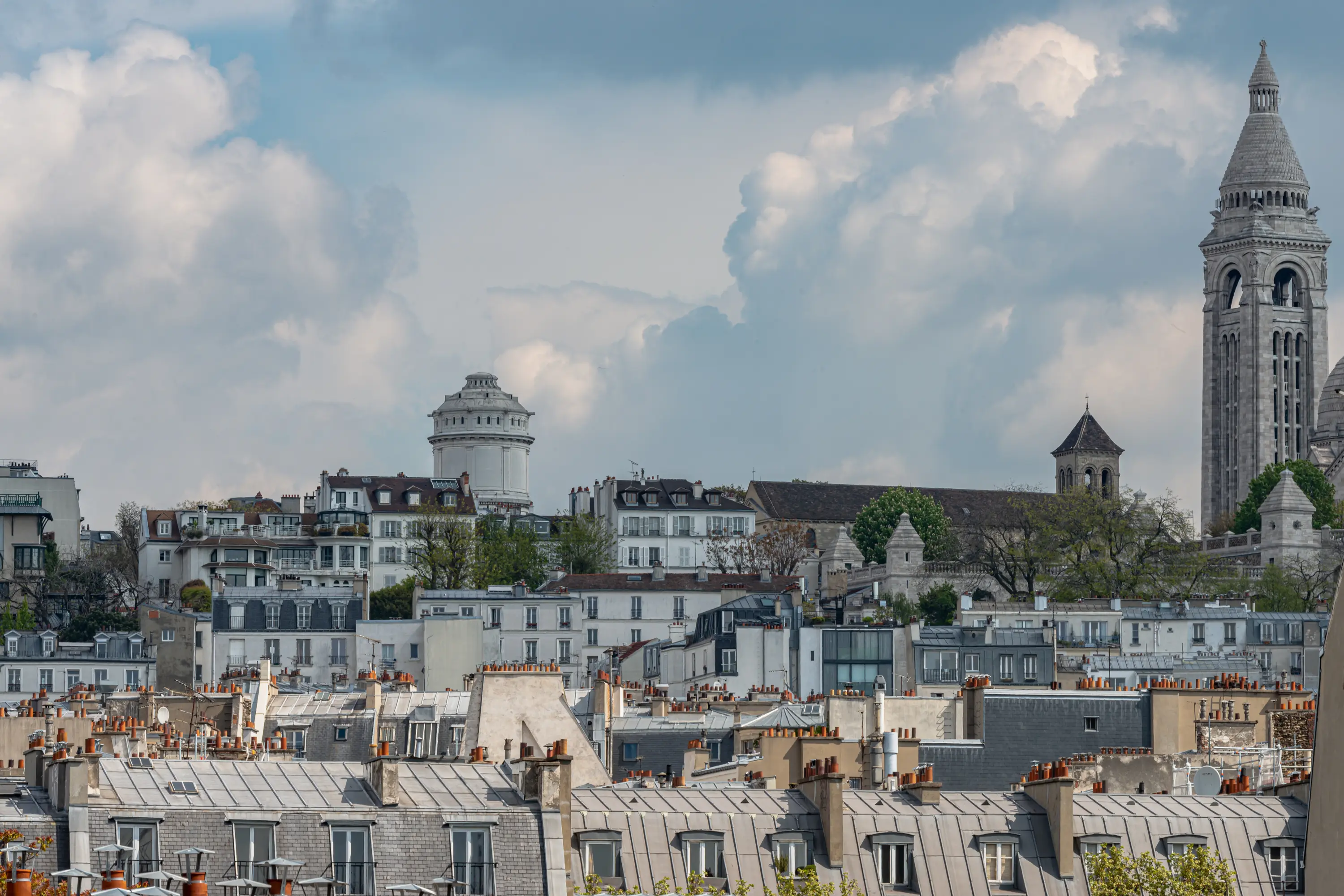 Paesaggio urbano di Parigi con tetti, alberi e la torre della Basilica del Sacro Cuore sotto un cielo nuvoloso.
