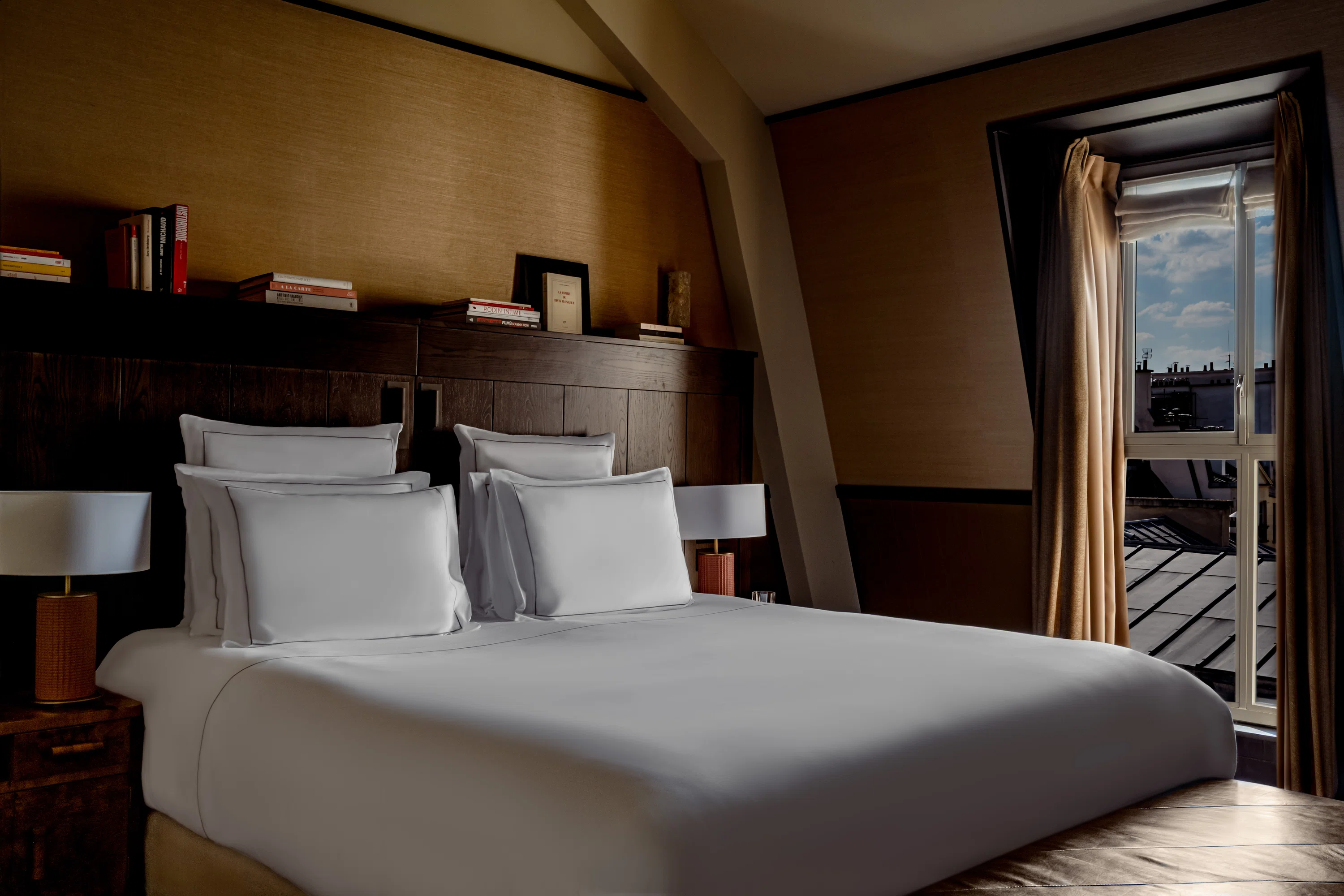 Habitación de hotel con una cama grande, ropa de cama blanca, lámparas de noche, cabecera de madera y ventana con vista a la ciudad.
