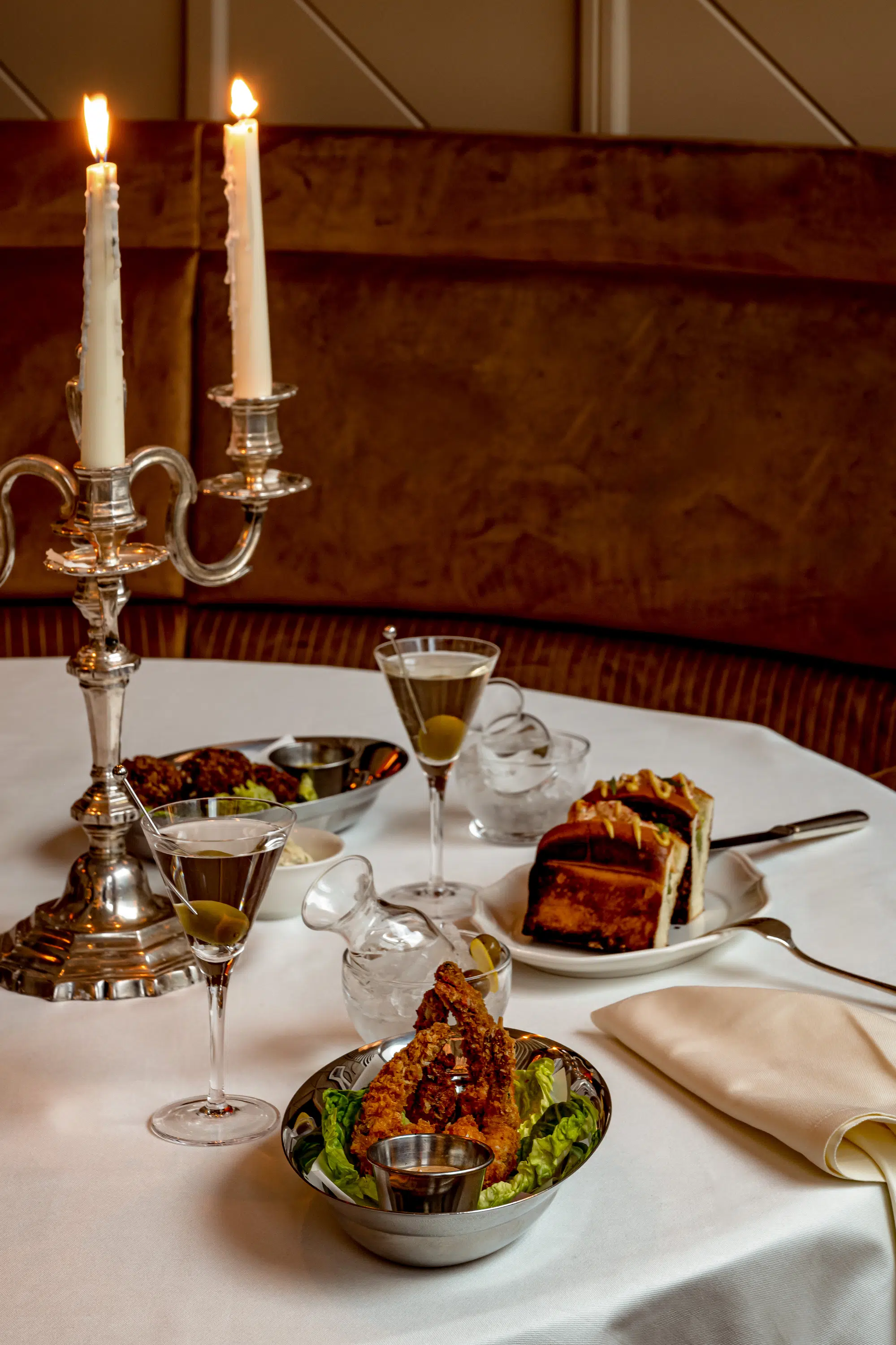 Mesa con dos martinis, pollo frito, sándwich, condimentos y velas encendidas en un soporte plateado.