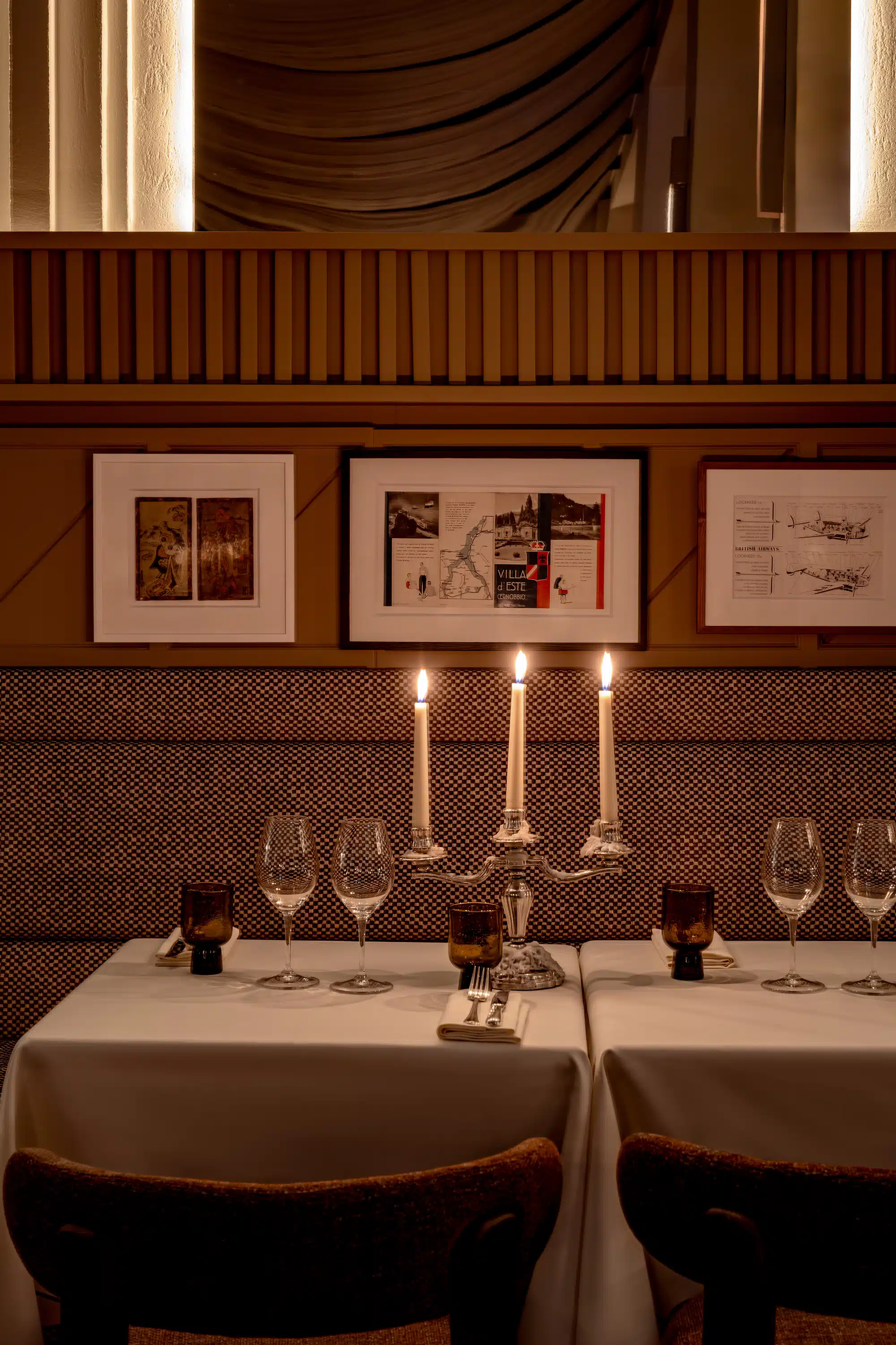Mesa de restaurante a la luz de las velas con vasos, sillas marrones y arte enmarcado en una pared estampada.