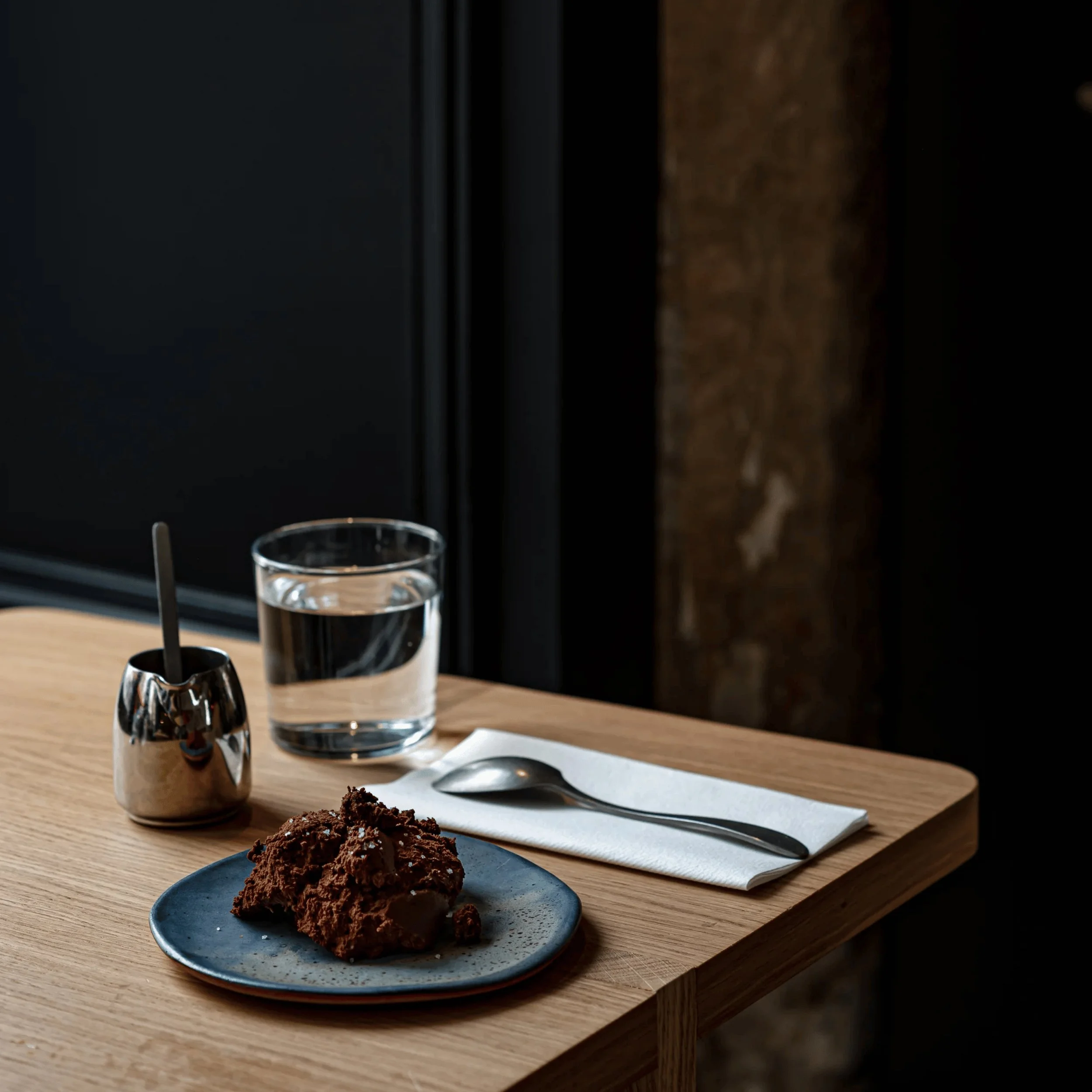 Dessert al cioccolato su piatto blu con cucchiaio, tovagliolo, bicchiere d'acqua e piccolo contenitore di metallo su tavolo di legno.
