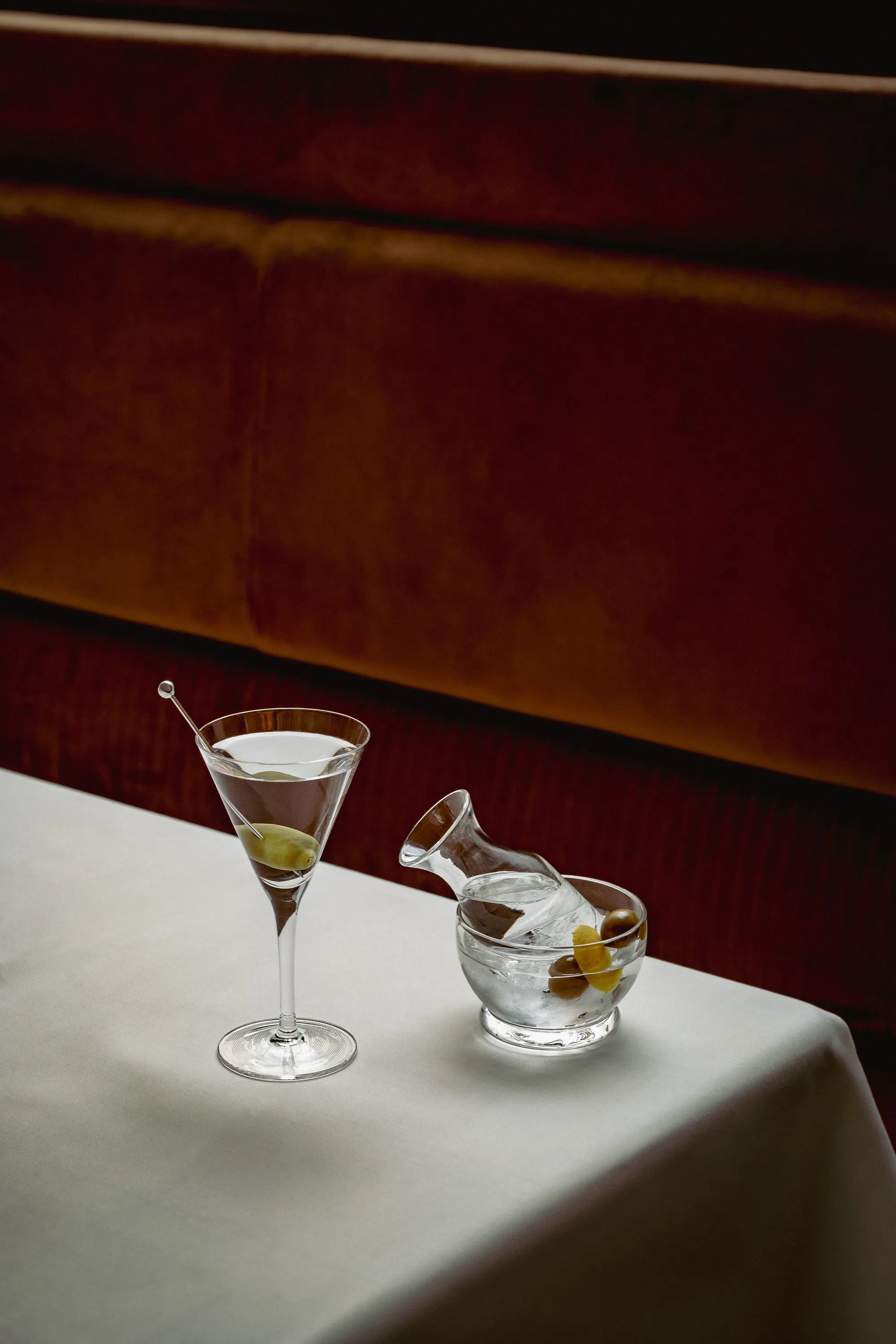 Copa de martini con aceituna y una jarra pequeña en un bol de cristal con aceitunas y limón sobre un mantel blanco.