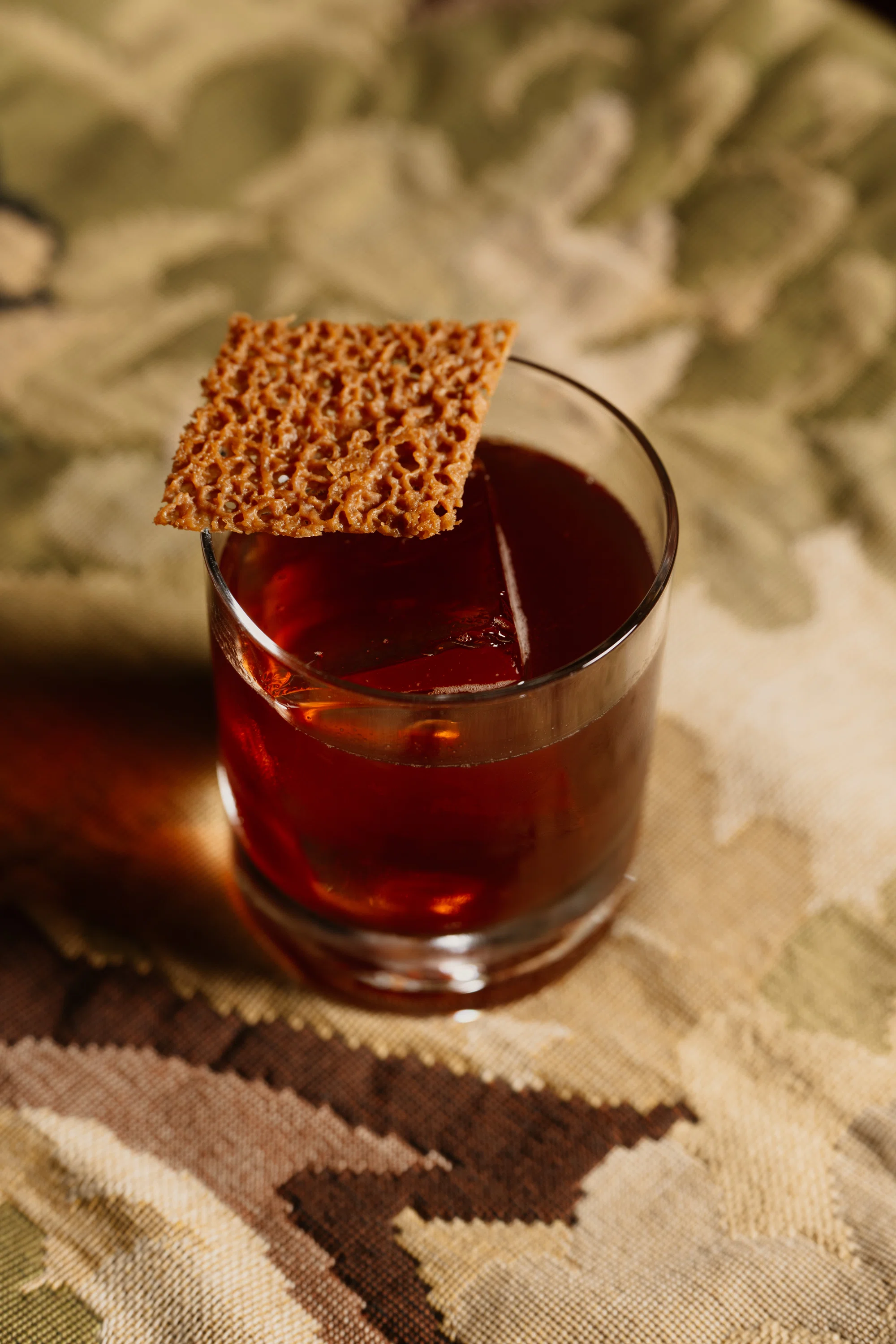 Vaso corto con cóctel rojo oscuro, cubitos de hielo grandes y guarnición cuadrada y crujiente en la parte superior.