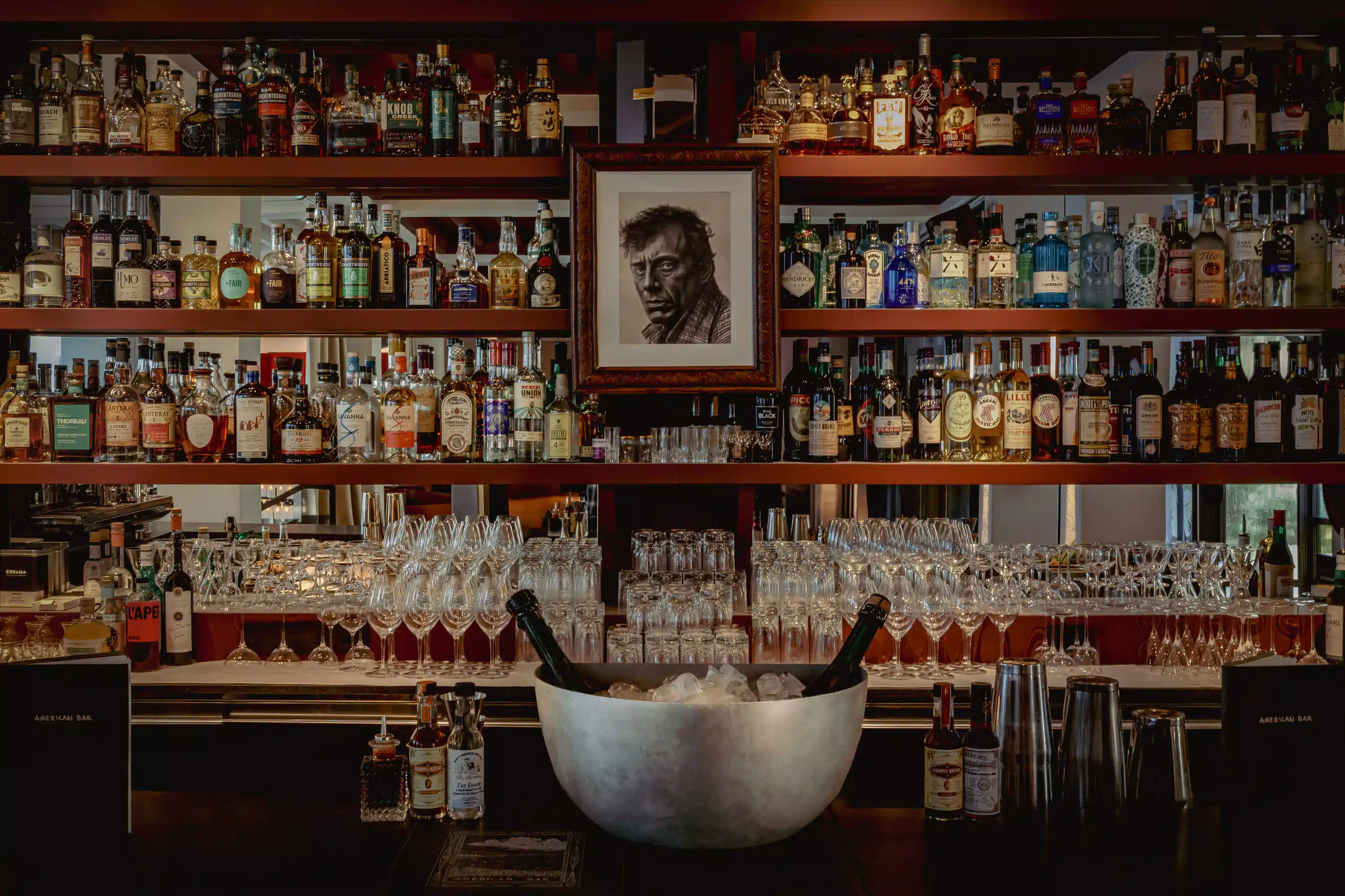 Los estantes del bar están llenos de botellas de licor y vasos vacíos detrás de un mostrador con un cubo de hielo y herramientas de bar.