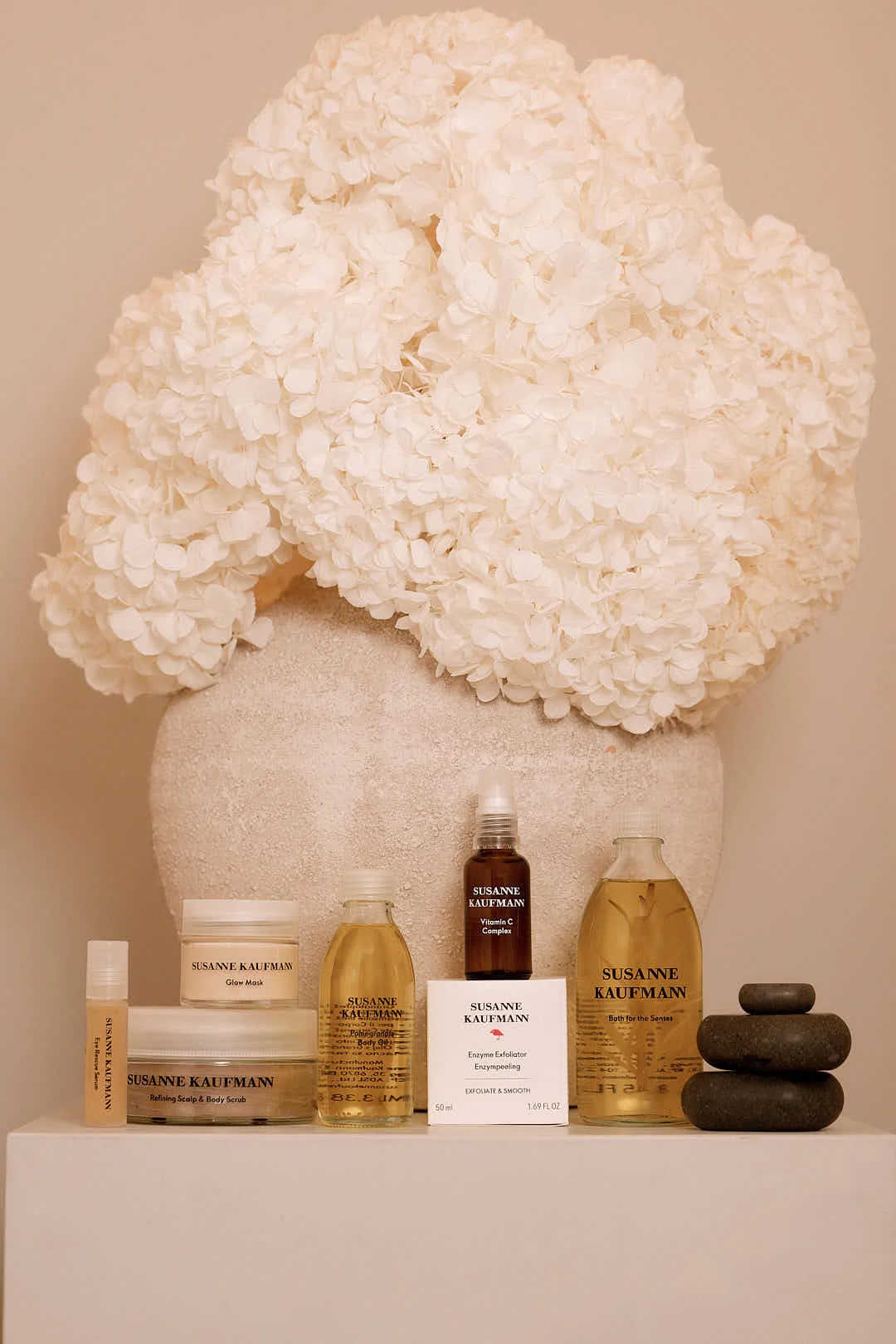 Los productos para el cuidado de la piel de Susanne Kaufmann se muestran con flores blancas y piedras de spa negras apiladas.
