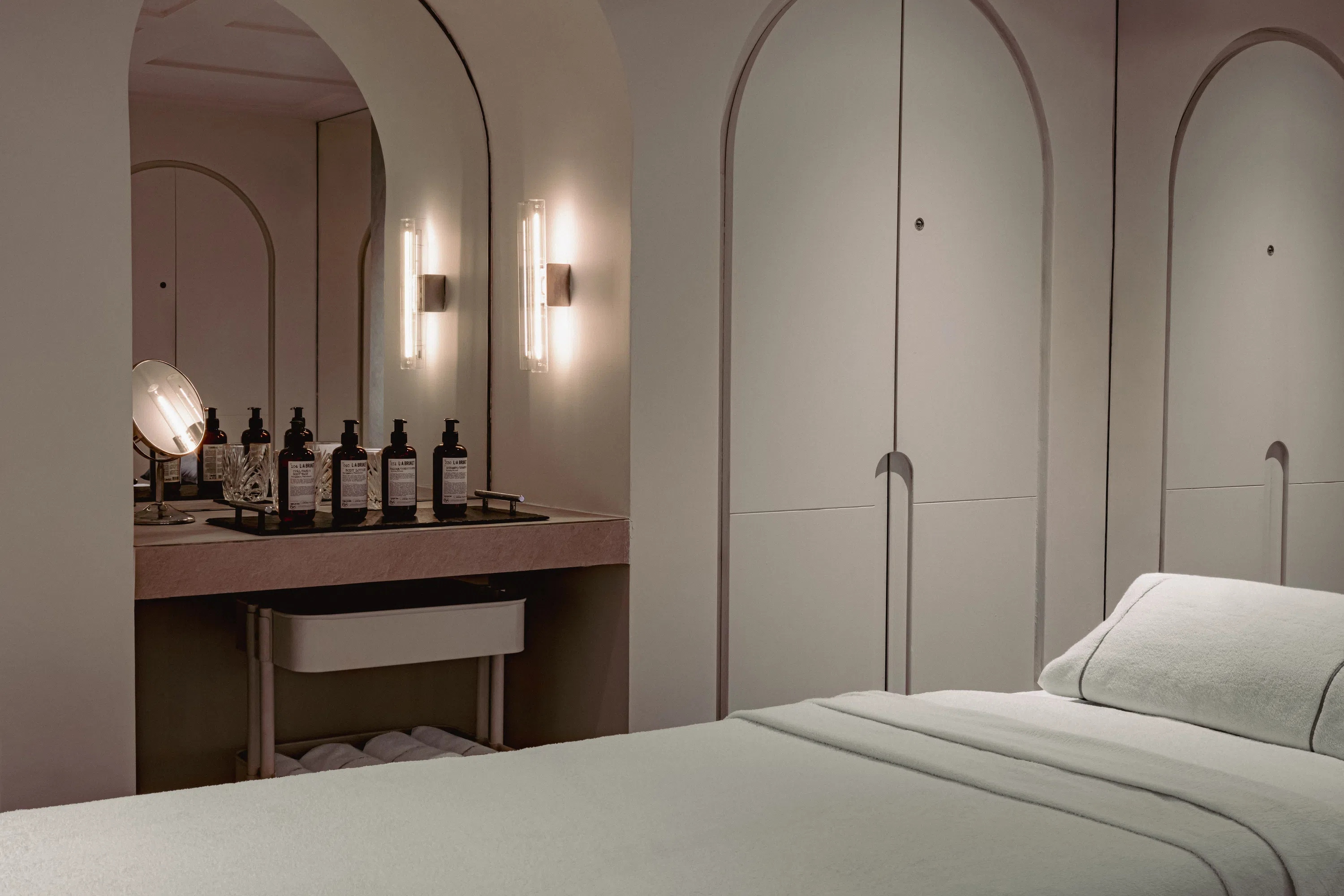Sala de spa minimalista con cama de tratamiento, iluminación tenue y botellas para el cuidado de la piel en un tocador con espejo.