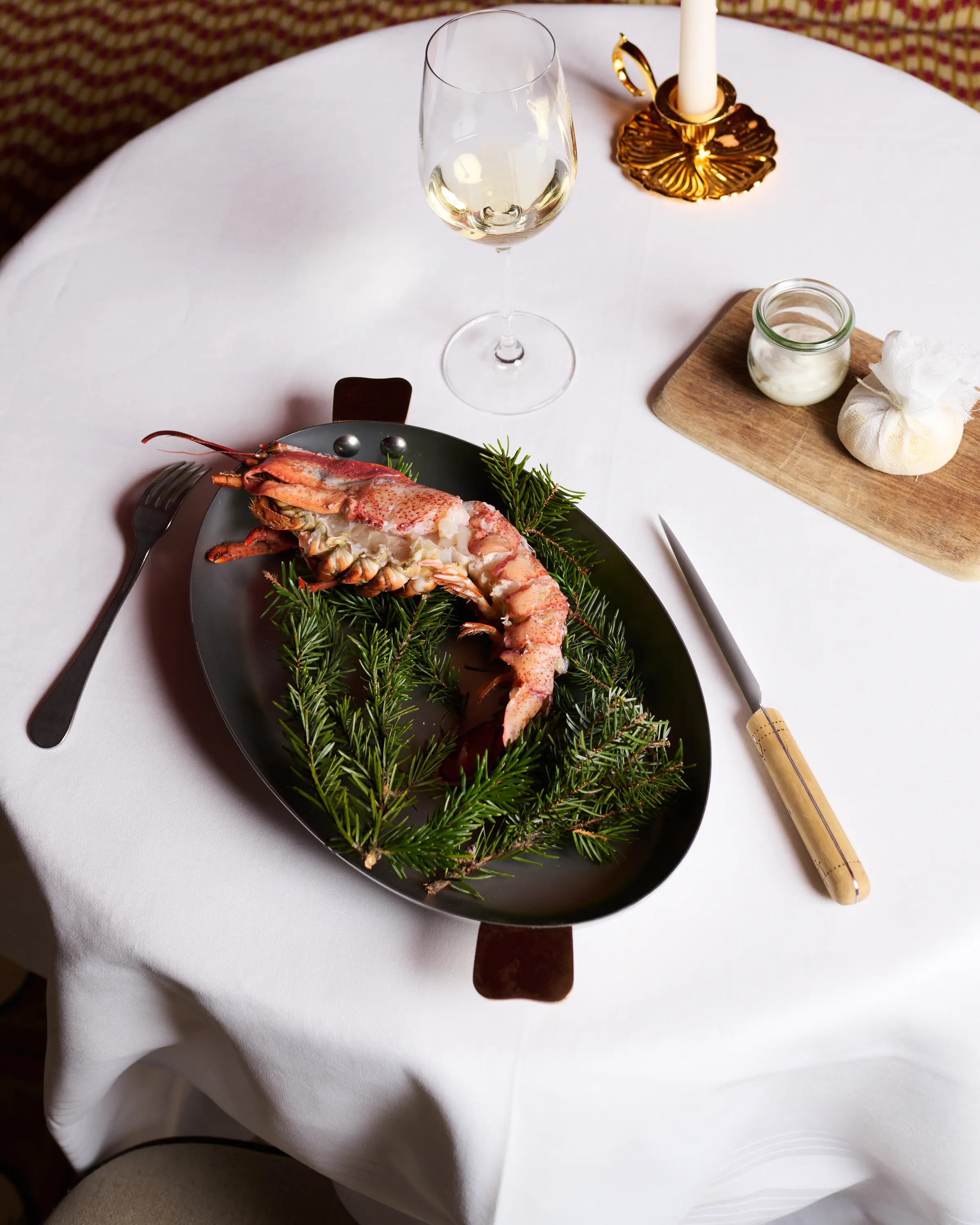 Coda di aragosta su piatto nero con rametti di pino verde, un bicchiere di vino bianco, forchetta, coltello e candela su tovaglia bianca.