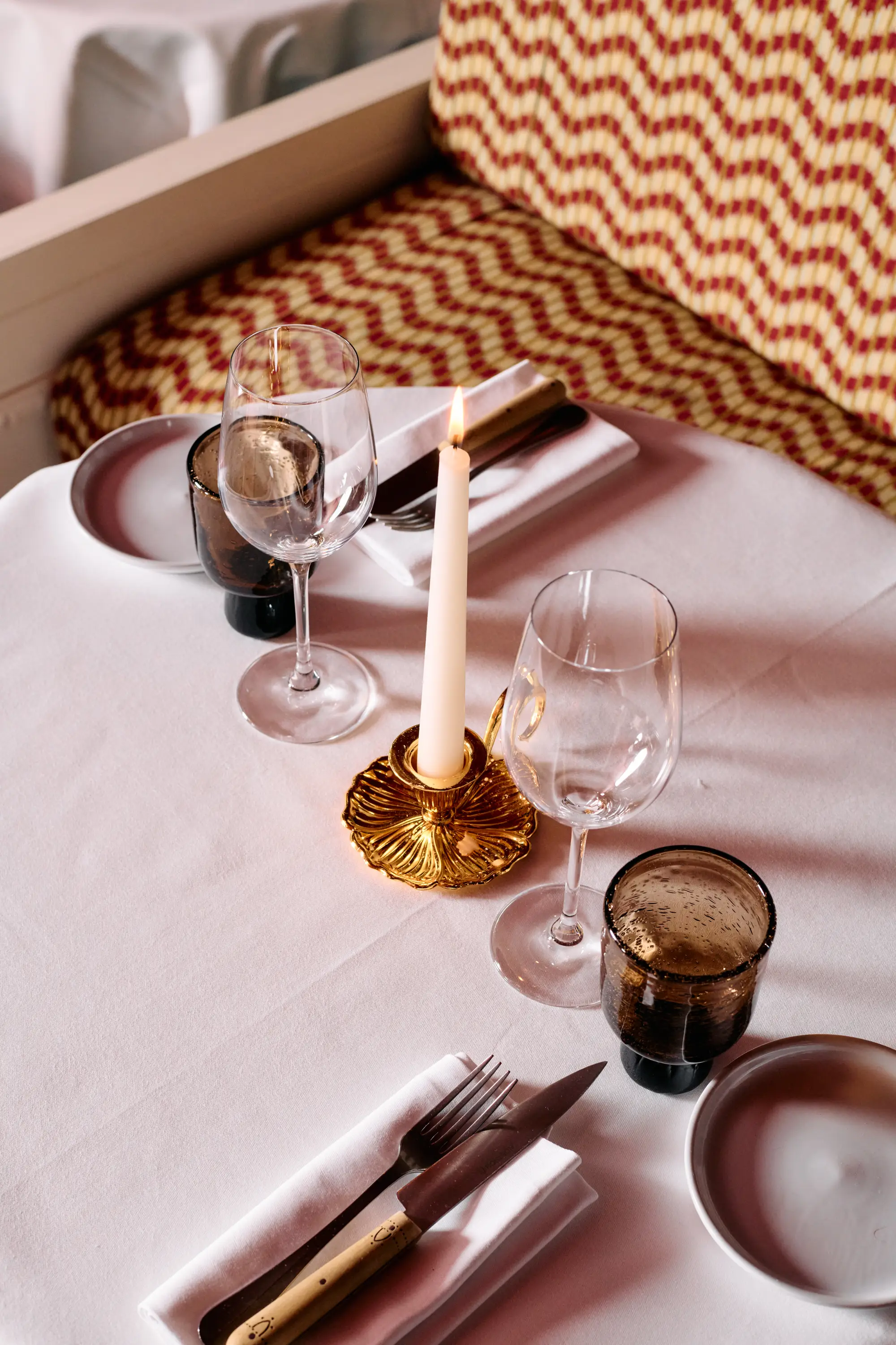 Table set with a lit candle, wine glasses, brown tumblers, white napkins, and cutlery near a patterned bench.