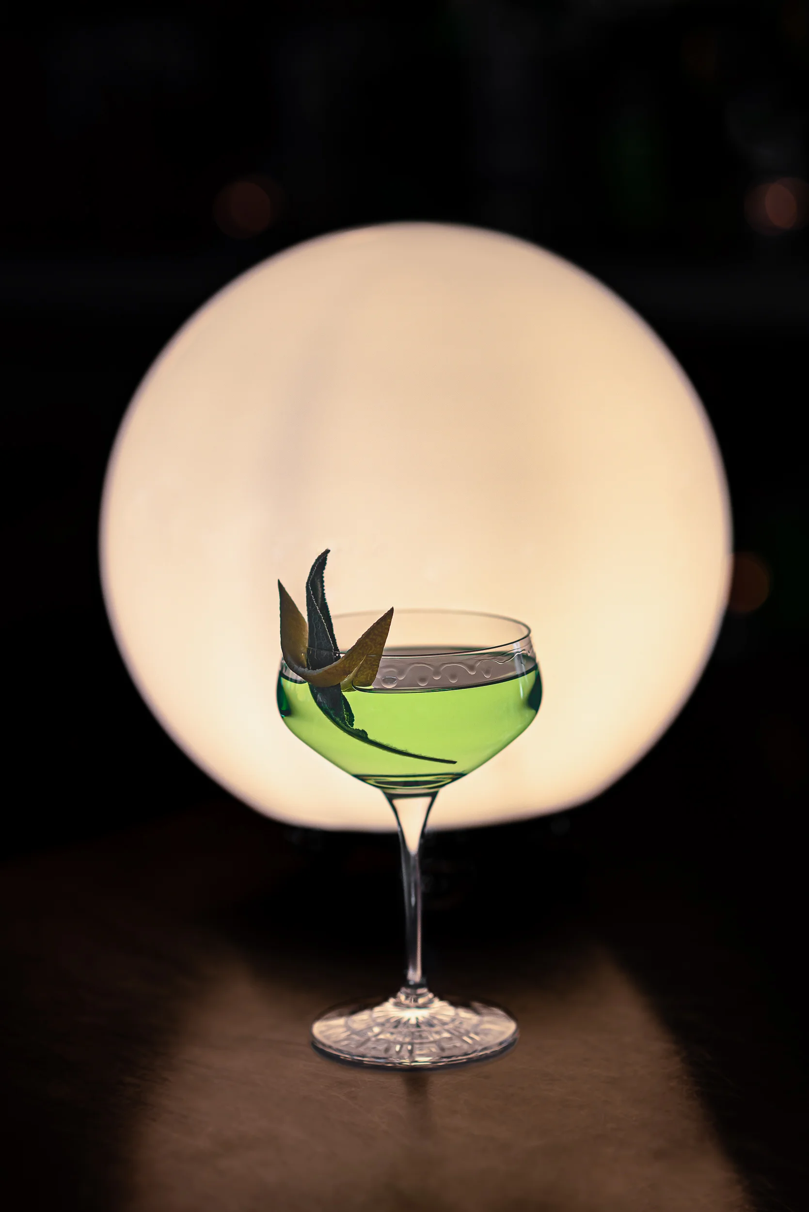 Elegante bicchiere da coupé con cocktail verde e contorno a foglie su un grande sfondo a sfera luminosa.