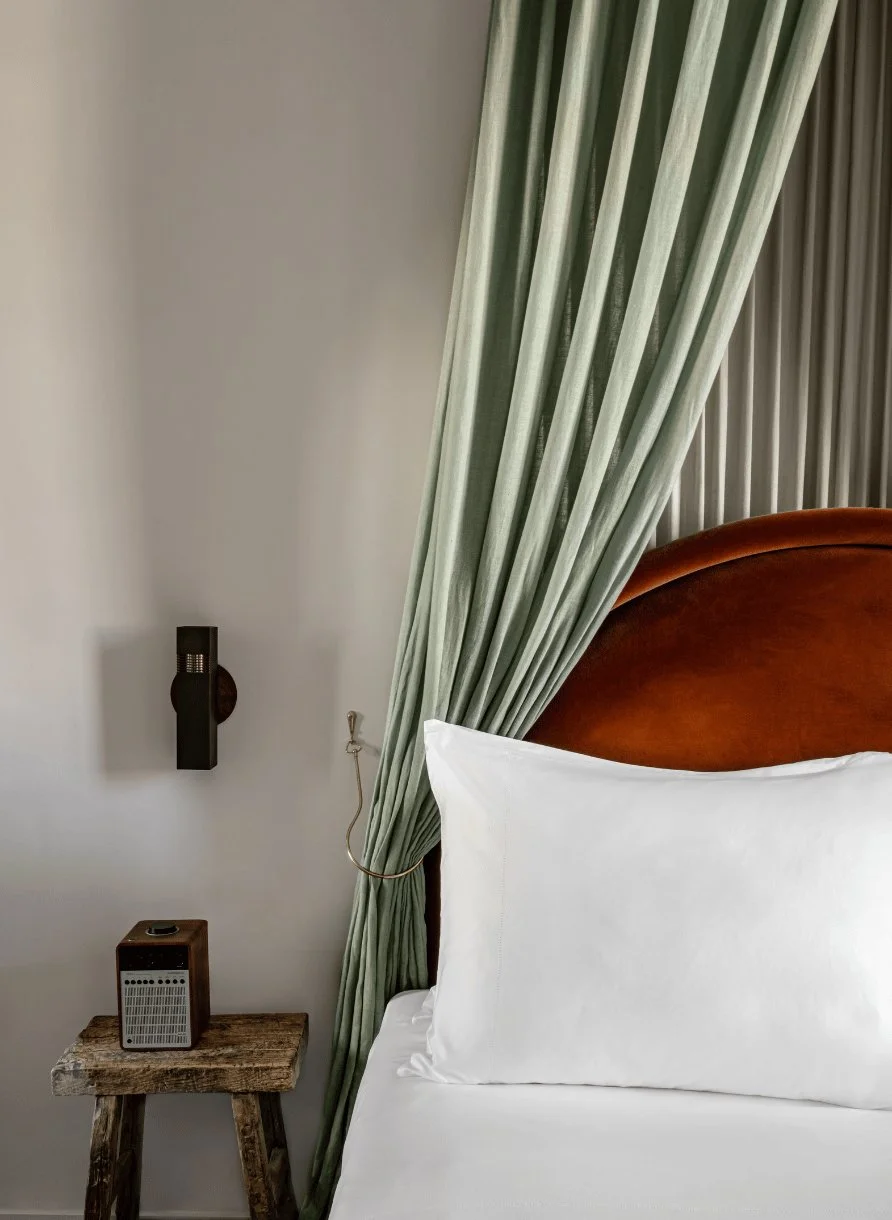 Almohada blanca sobre una cama con cabecero marrón curvo, cortina verde, mesa auxiliar de madera rústica y lámpara de pared