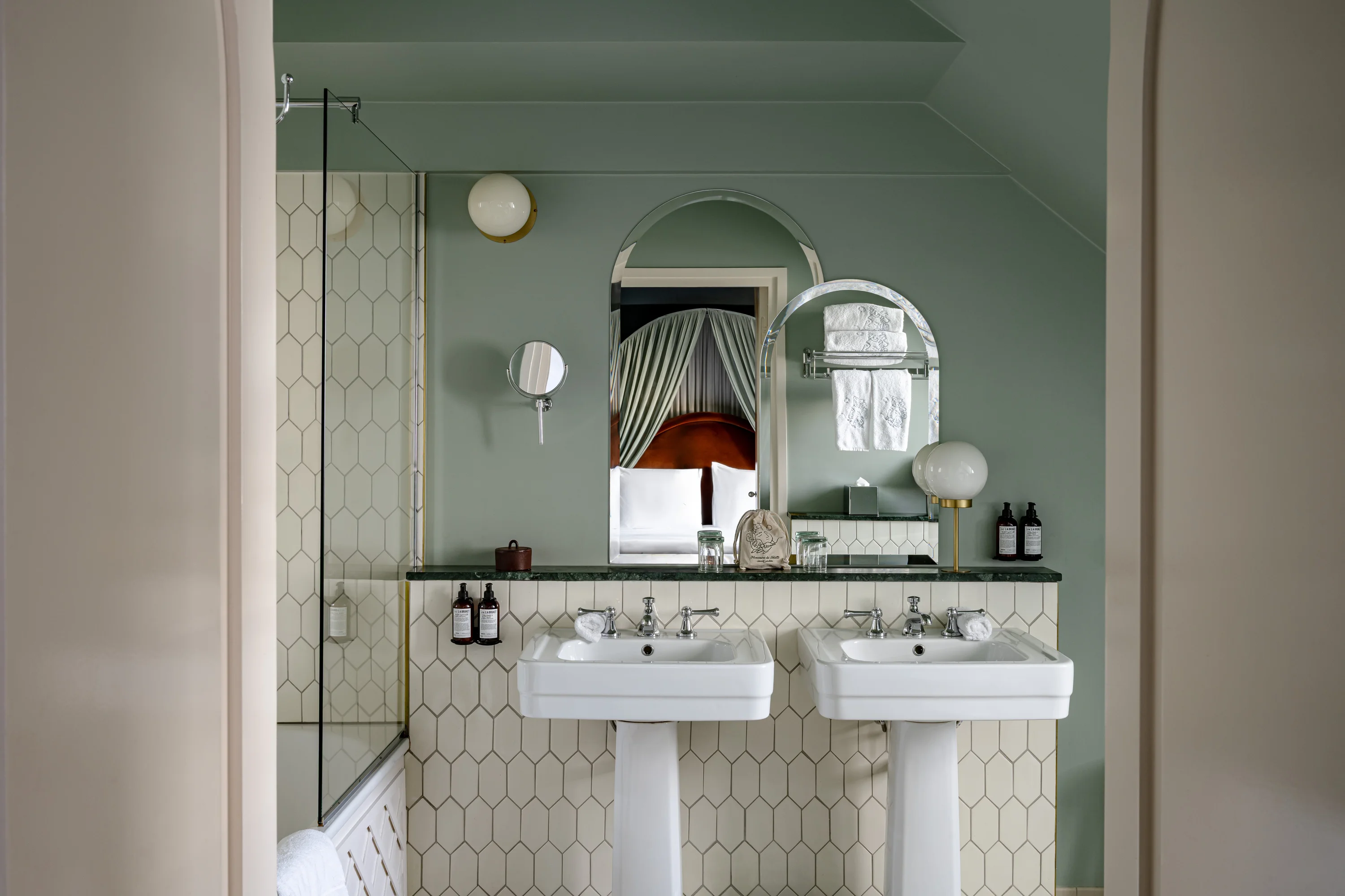 Baño con dos lavabos blancos con pedestal, paredes verdes, azulejos hexagonales y un espejo que refleja una cama.