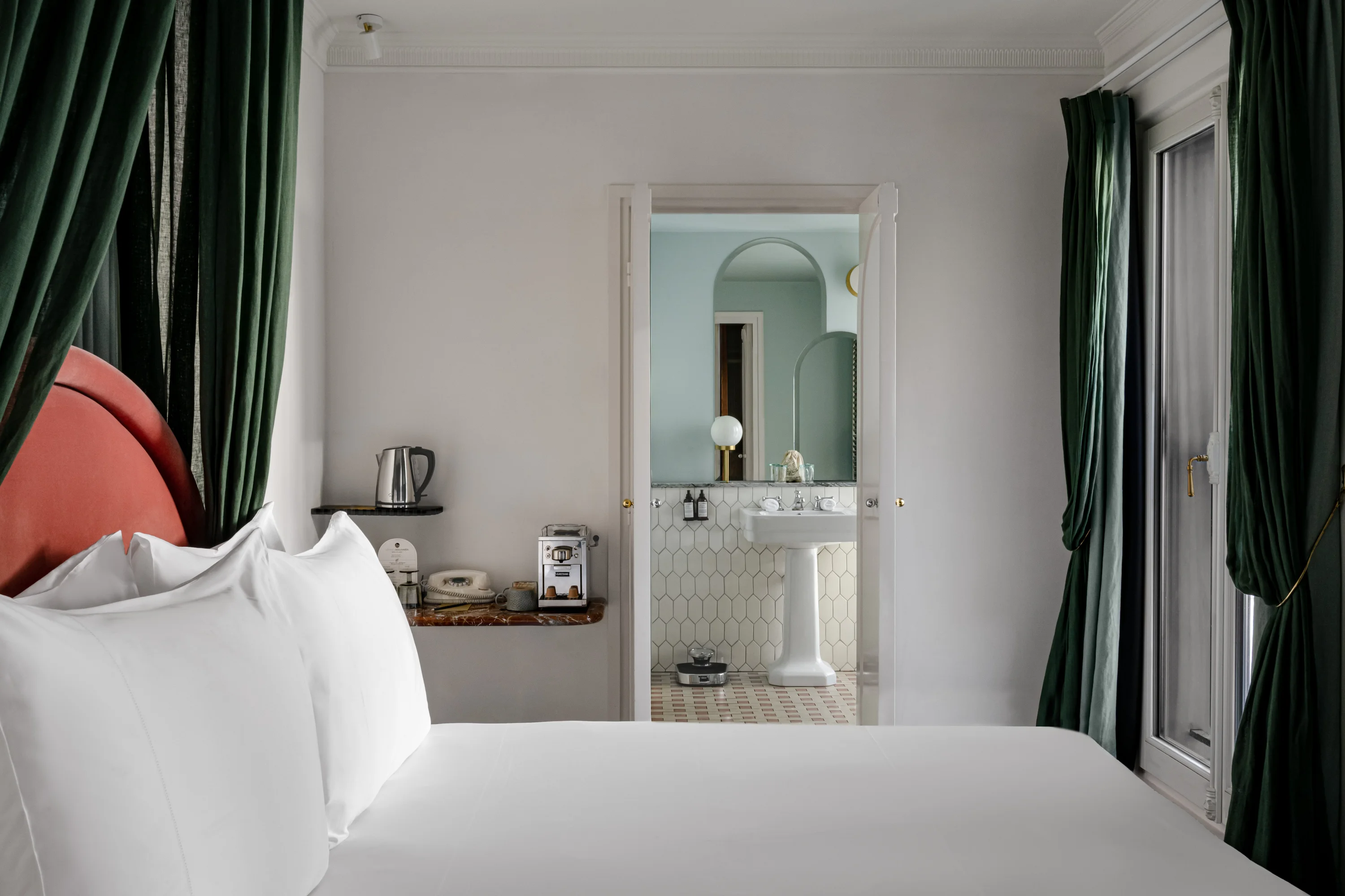 Habitación de hotel con cama blanca, cortinas verdes y una puerta abierta que muestra un lavabo y un espejo.
