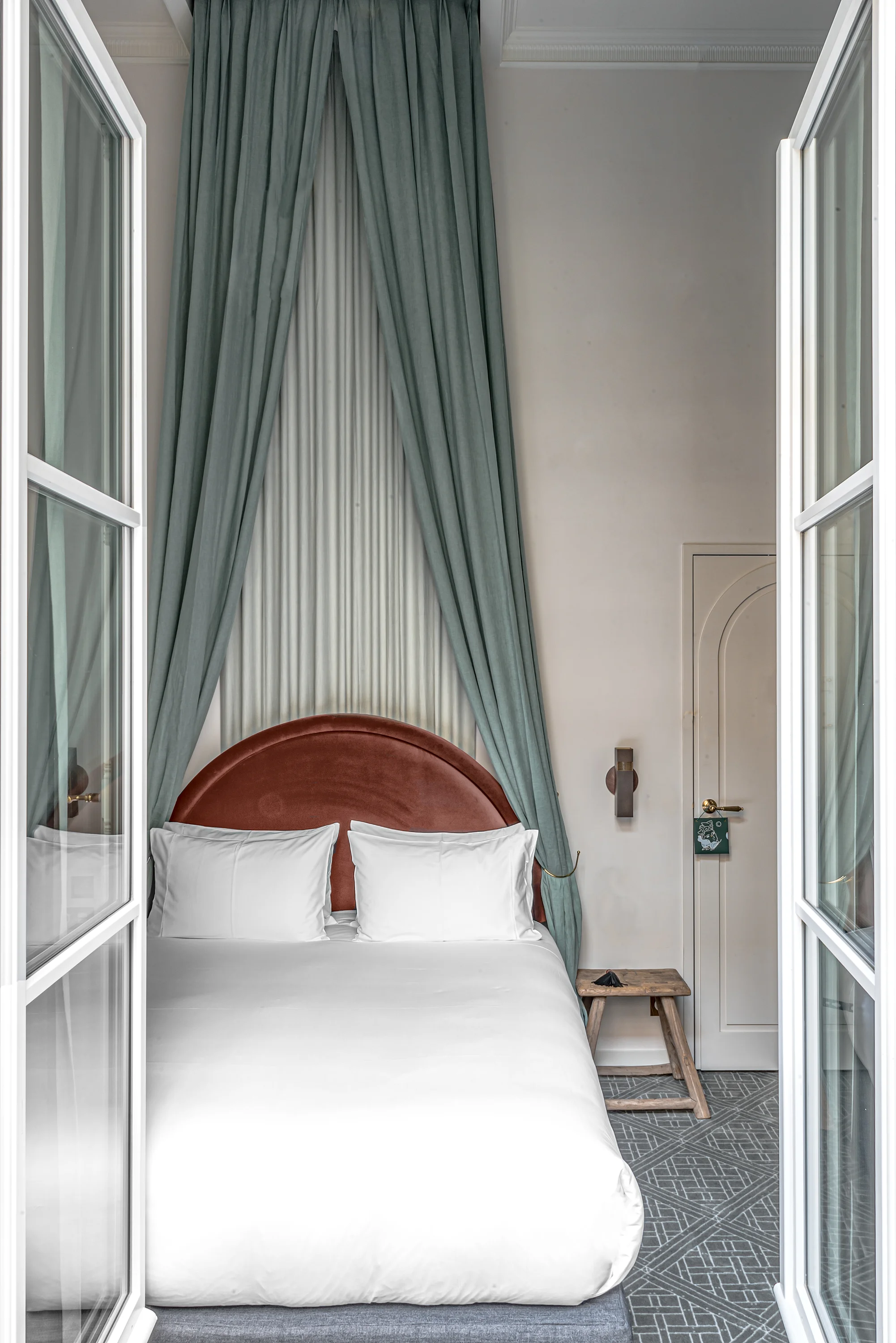 Bed with curved brown headboard, white bedding, flanked by green drapes and open glass doors.