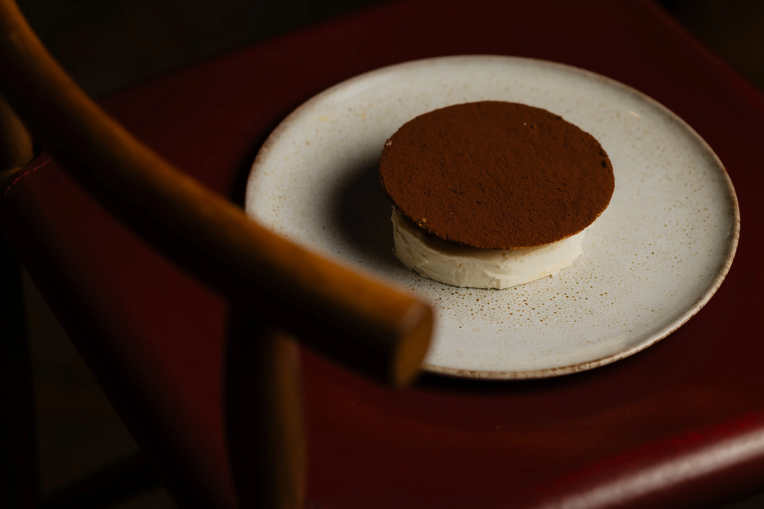 Postre redondo de chocolate con cacao en polvo sobre placa blanca sobre superficie de cuero rojo.