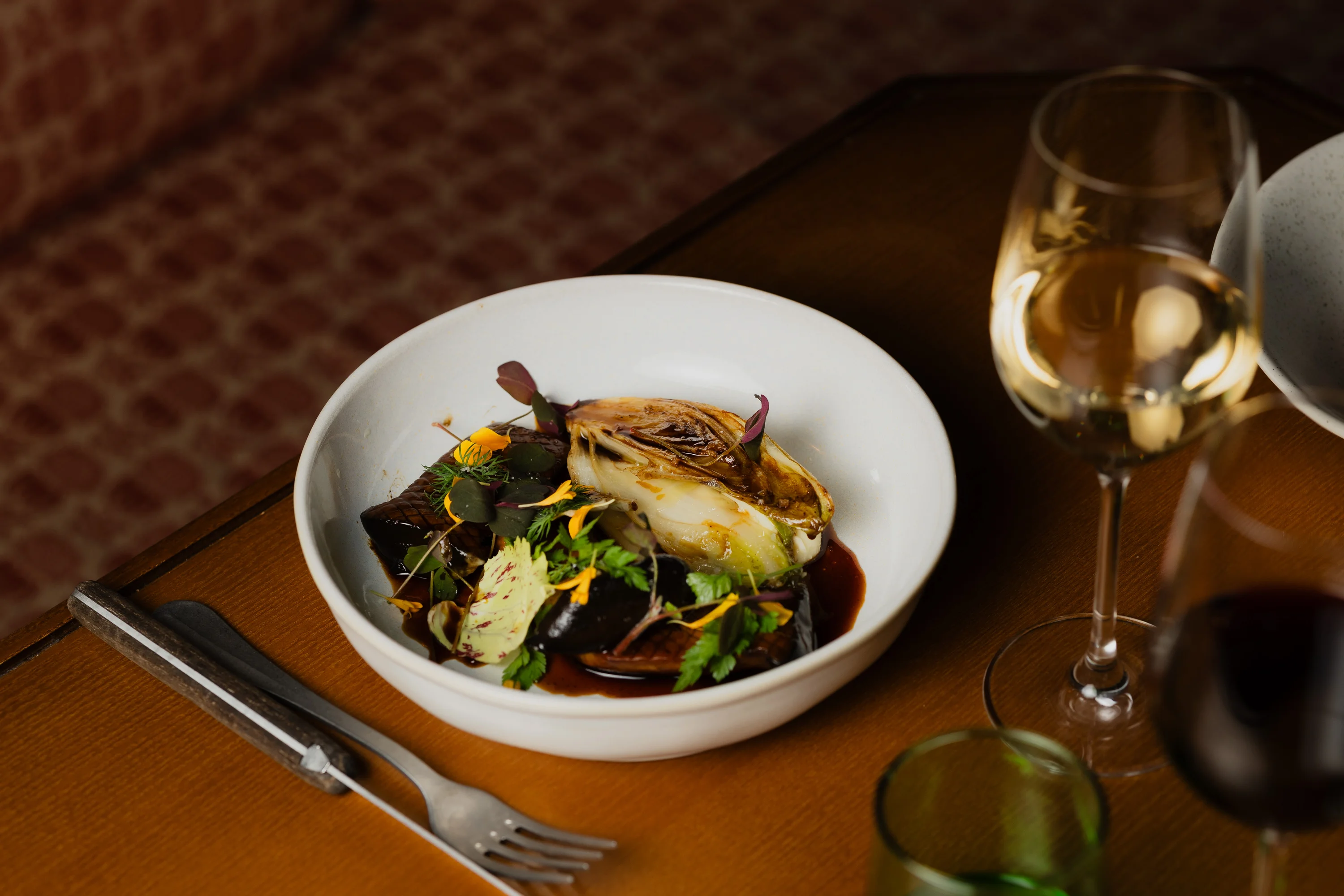 White bowl with roasted vegetables, greens, and edible flowers on a wooden table beside wine glasses
