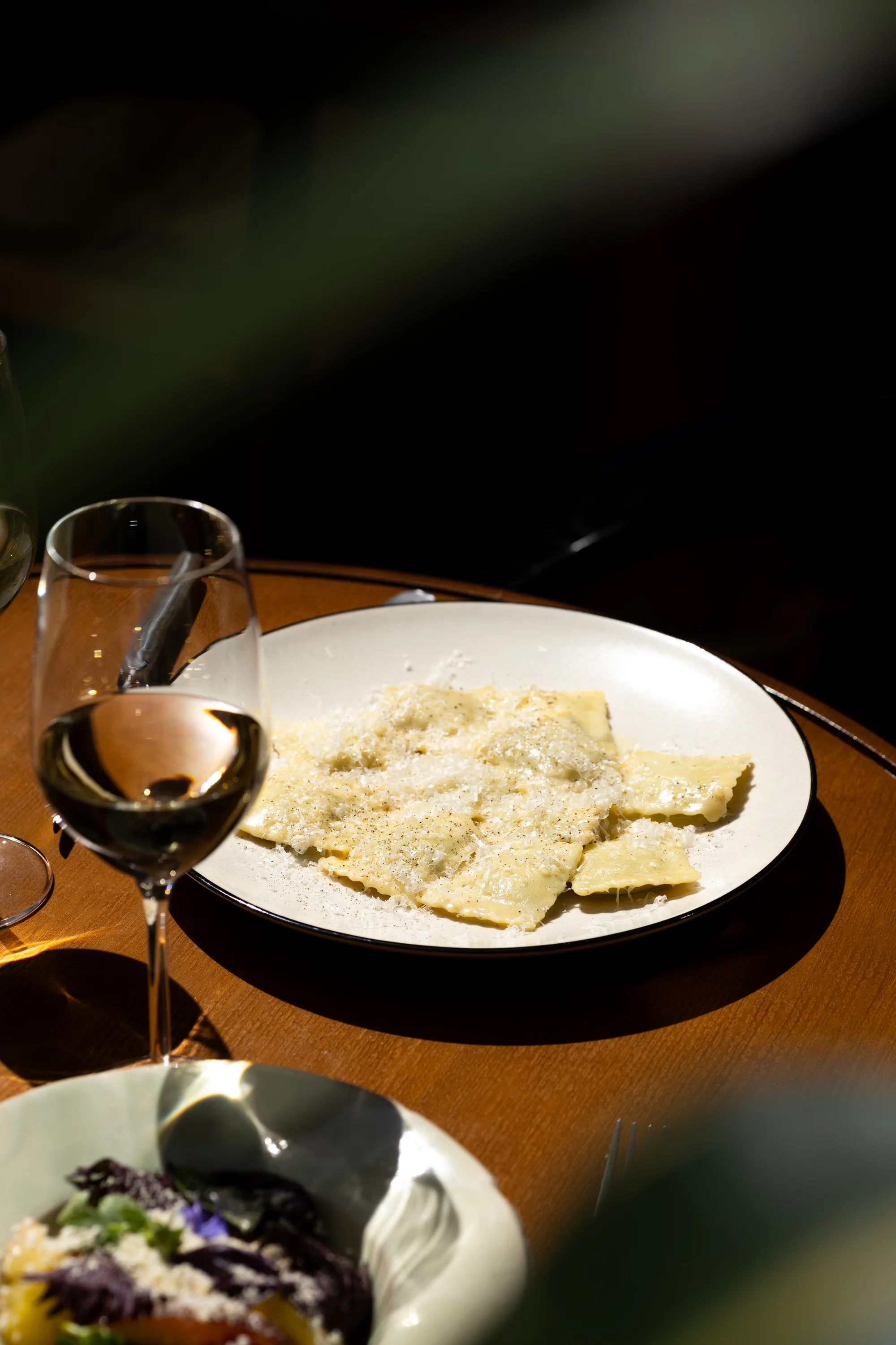 Plato de ravioles espolvoreado con queso rallado y una copa de vino blanco sobre una mesa de madera.