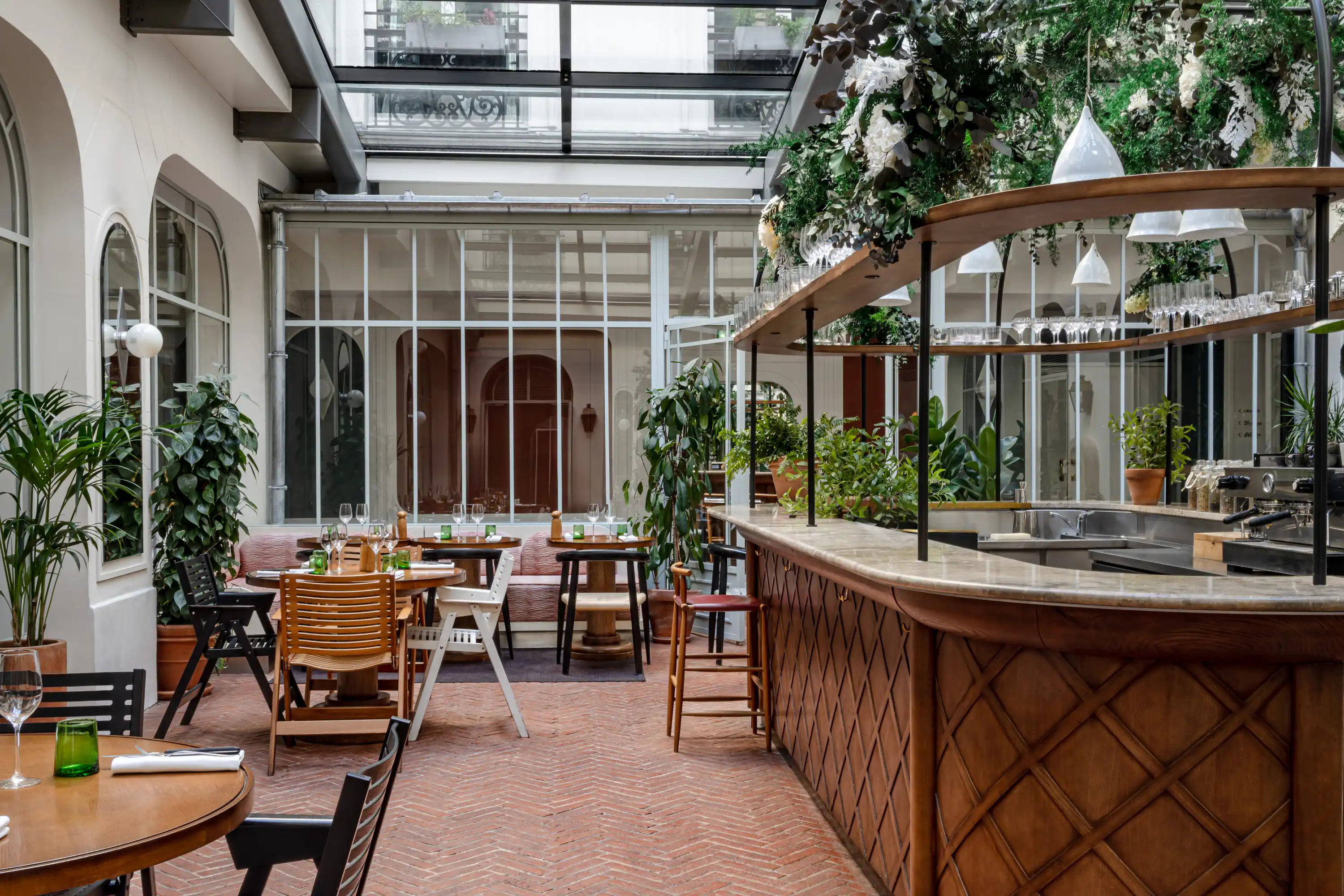 Elegante interior de restaurante con mesas de madera, sillas, zonas verdes y una barra curva bajo un techo de cristal.
