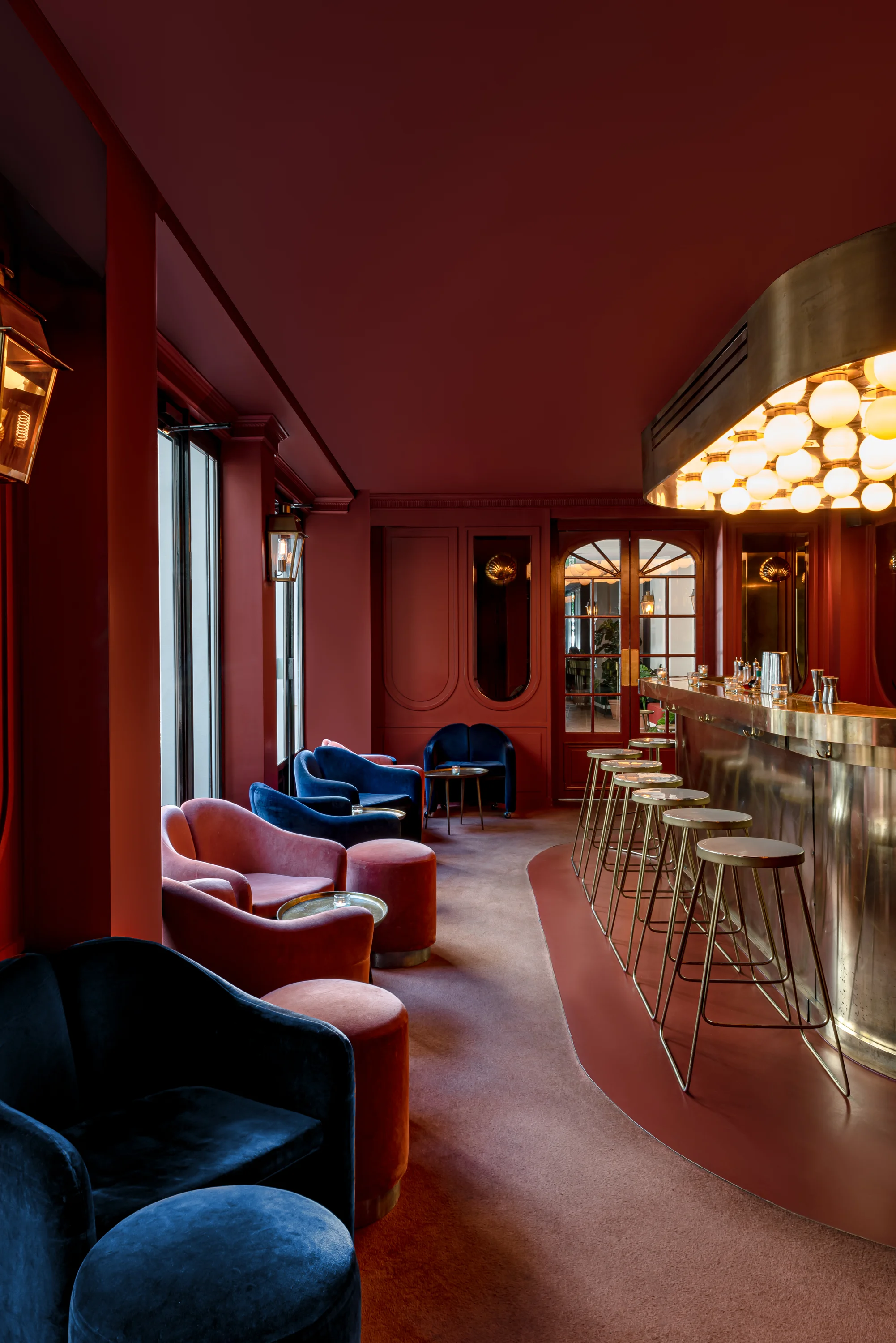 Elegante interior de bar con taburetes dorados, sillones de terciopelo en azul y rosa, paredes rojas e iluminación cálida.