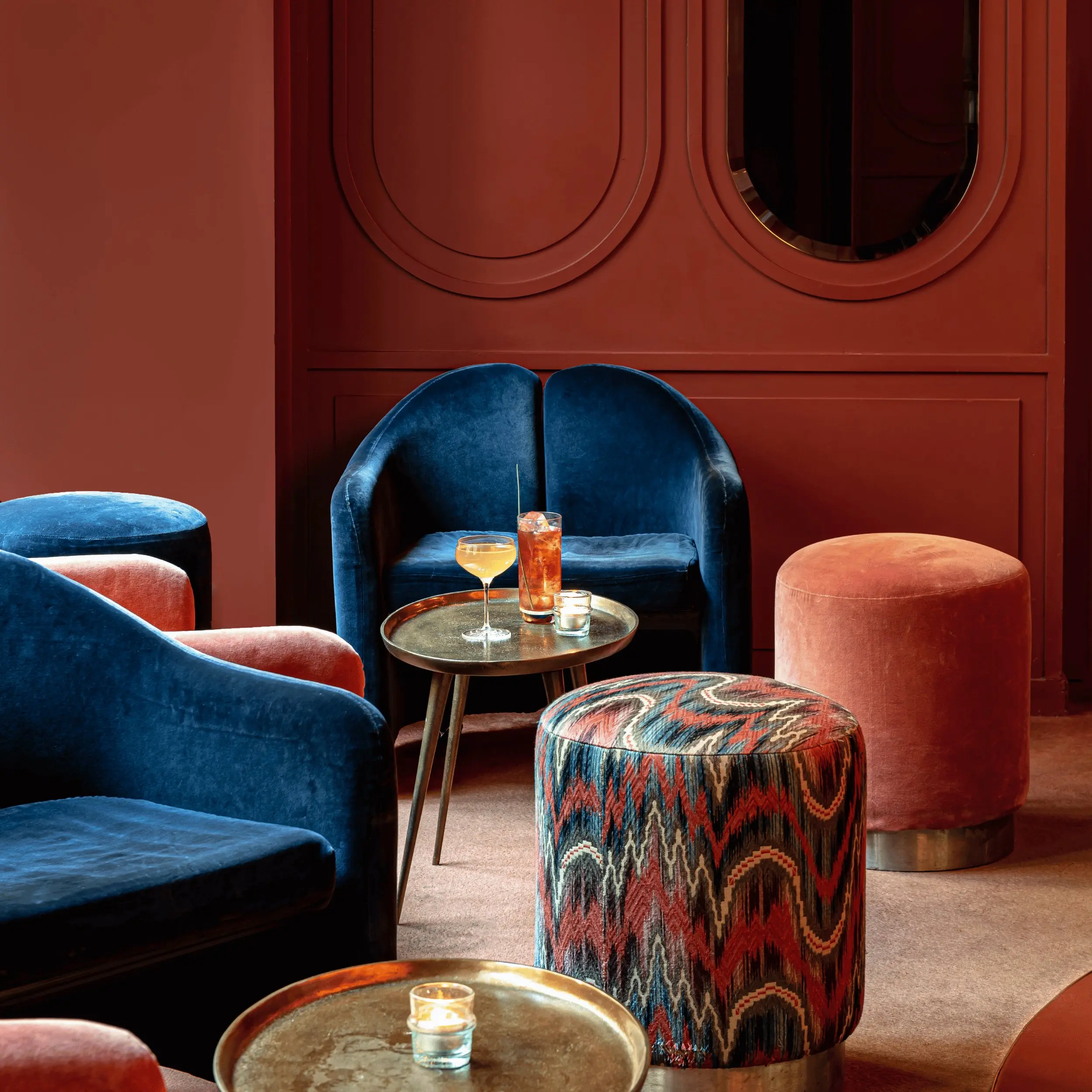 Acogedor salón con sillas de terciopelo azul, pufs estampados y rosas, pequeñas mesas redondas y una bebida en la mesa.