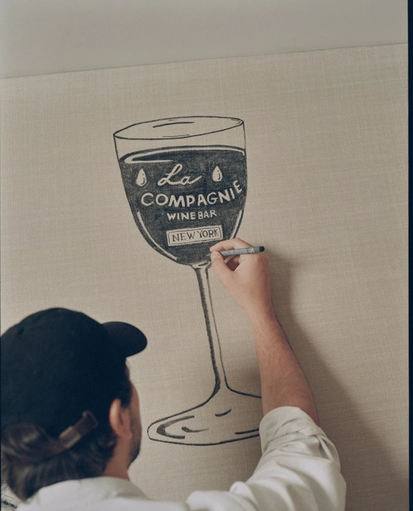 Pessoa desenhando o logotipo de uma taça de vinho com o texto La Compagnie Wine Bar New York em uma superfície bege.