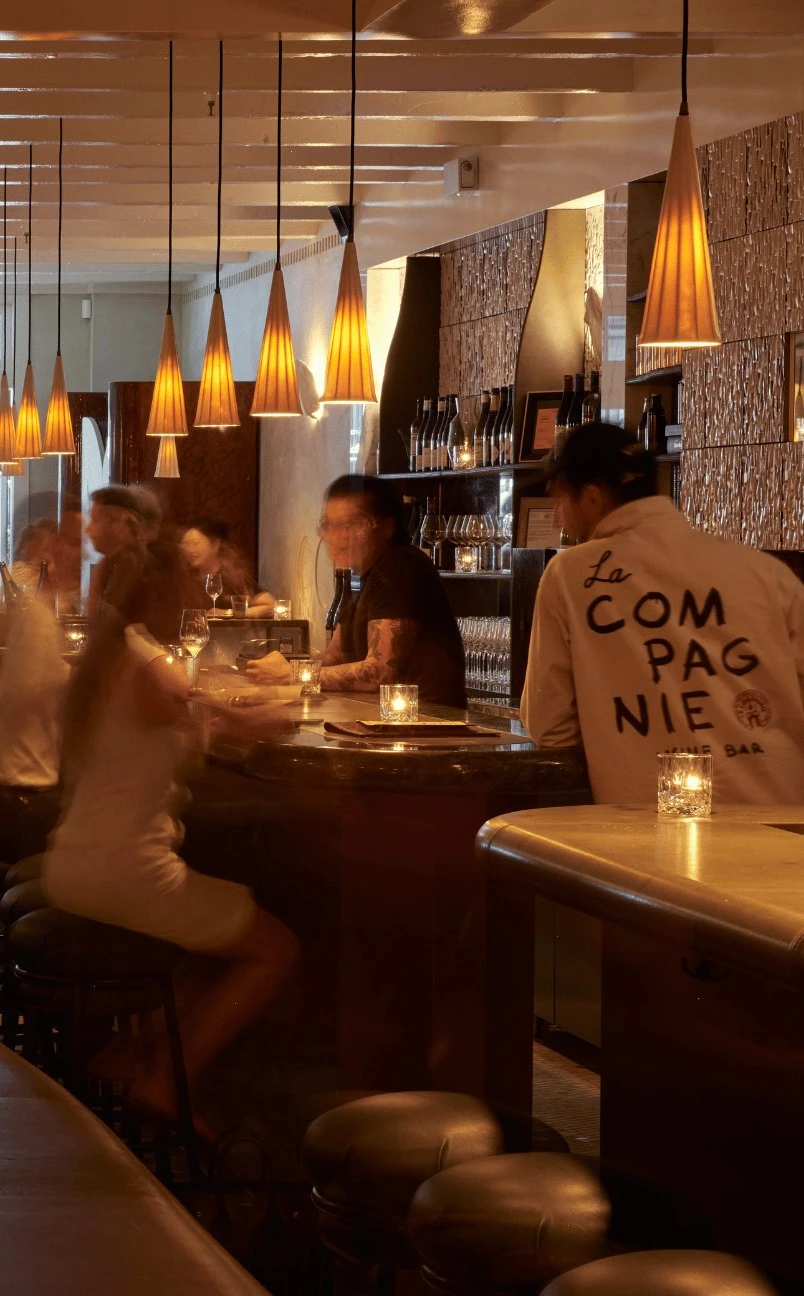 Bar mal iluminado com luzes penduradas, pessoas sentadas conversando no balcão com bebidas e velas.