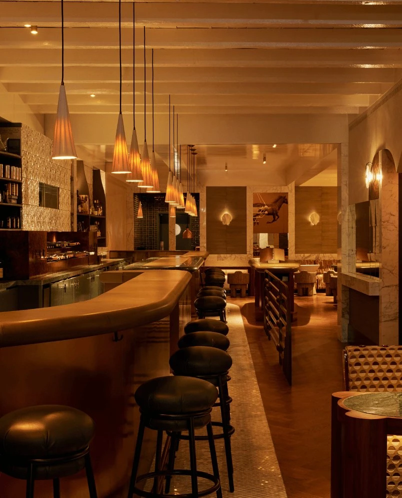 Bar moderno com iluminação aconchegante, balcão curvo, bancos pretos, luzes pendentes e assentos aconchegantes ao fundo.
