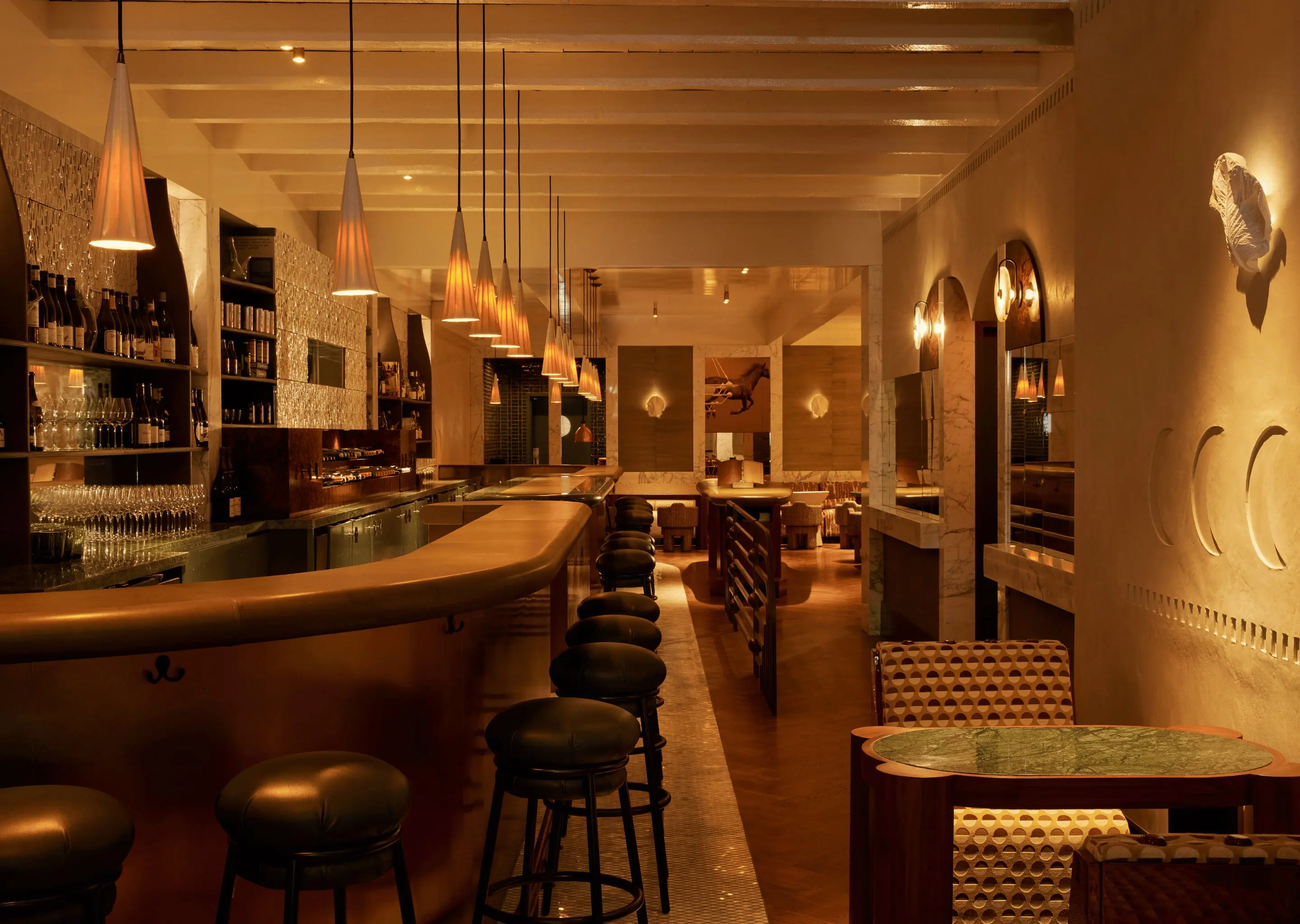 Bar de hotel com iluminação aconchegante, balcão de latão, luzes pendentes, bancos de couro e área de estar estampada ao fundo.