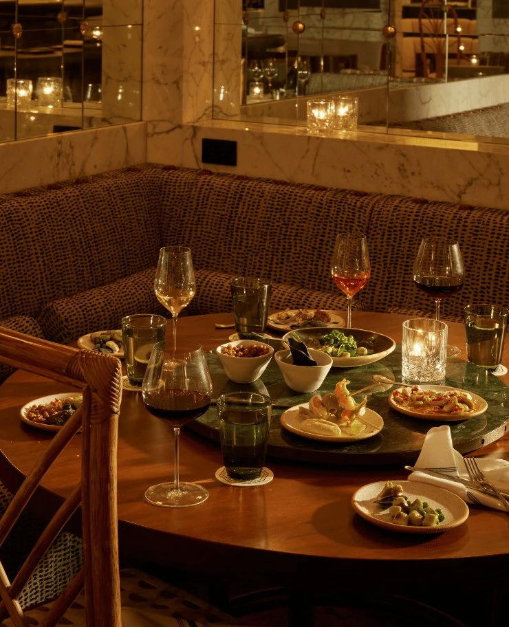 Mesa redonda com taças de vinho, pratos de aperitivos variados, copos de água verde e uma vela acesa.
