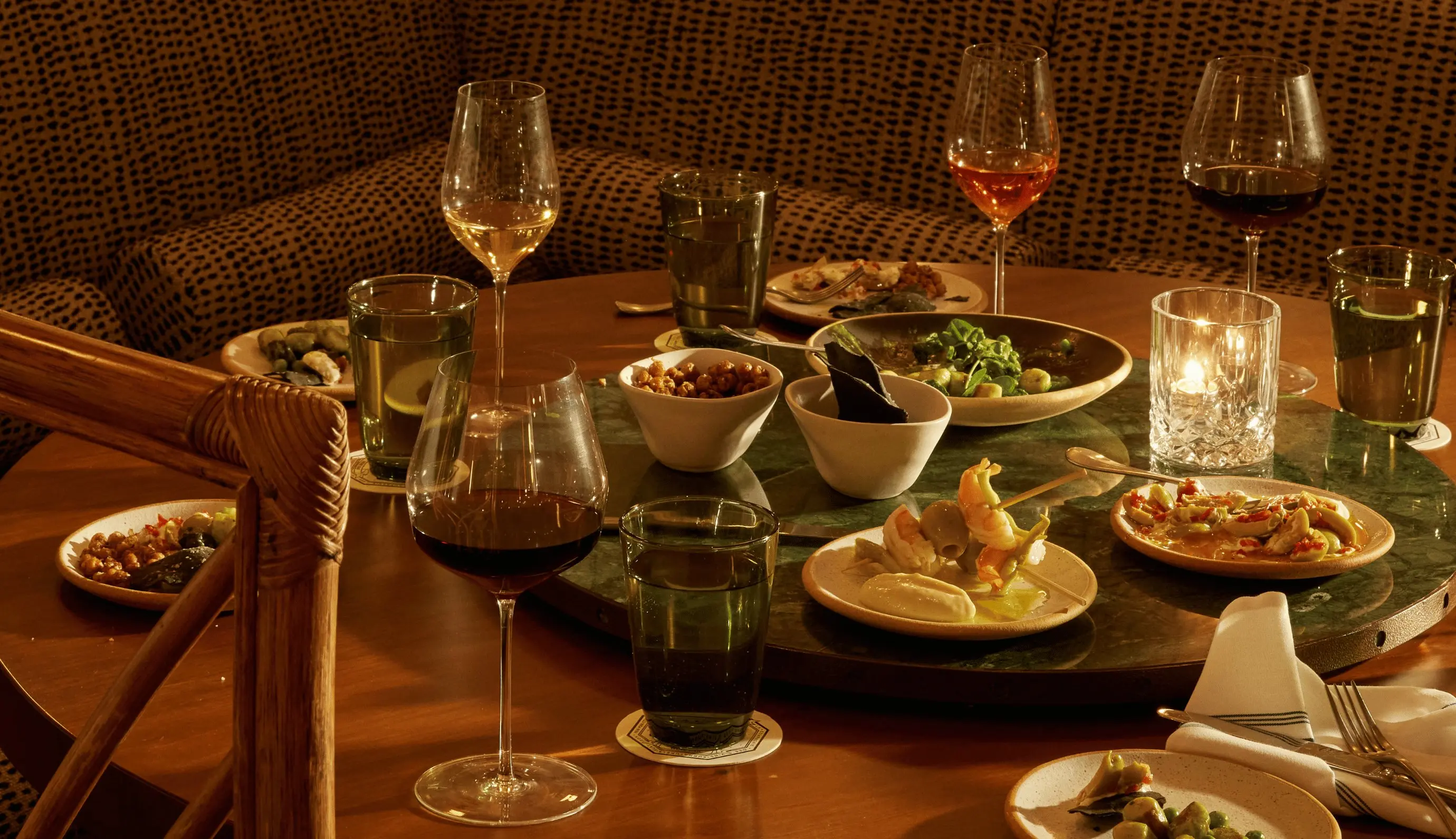 Mesa redonda com pratos variados, bebidas e taças de vinho em um ambiente de jantar bem iluminado.