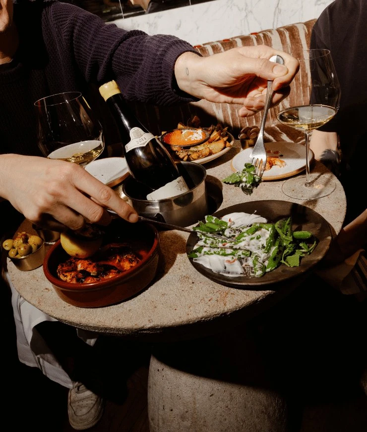 Duas pessoas em uma mesa com taças de vinho, uma garrafa em um refrigerador, azeitonas e pratos de comida.