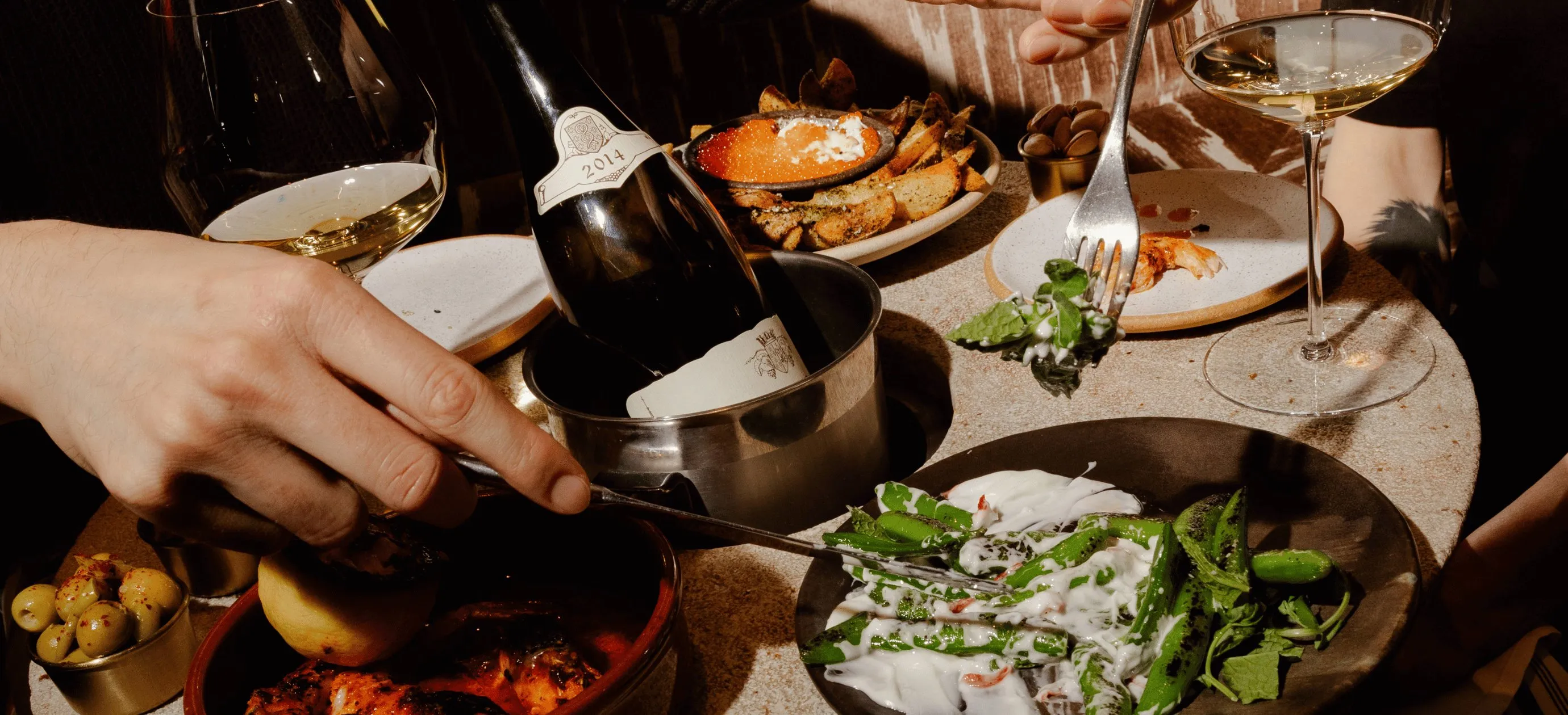 Duas pessoas compartilham uma refeição com vinho, azeitonas temperadas, batatas, vegetais grelhados e molho em uma mesa redonda.