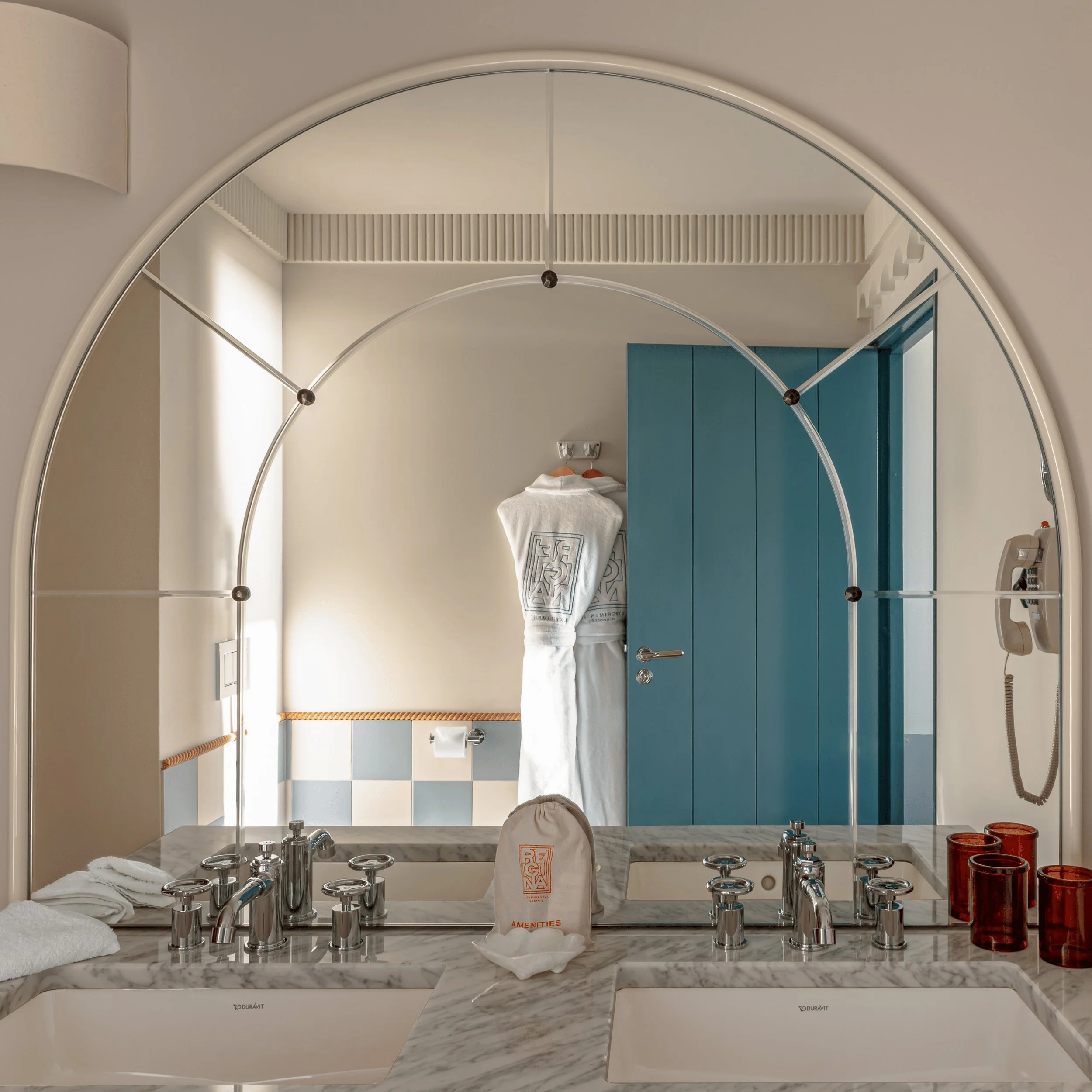 Bagno moderno con doppio lavabo in marmo, porta blu, accappatoio bianco e tazze in vetro ambrato.