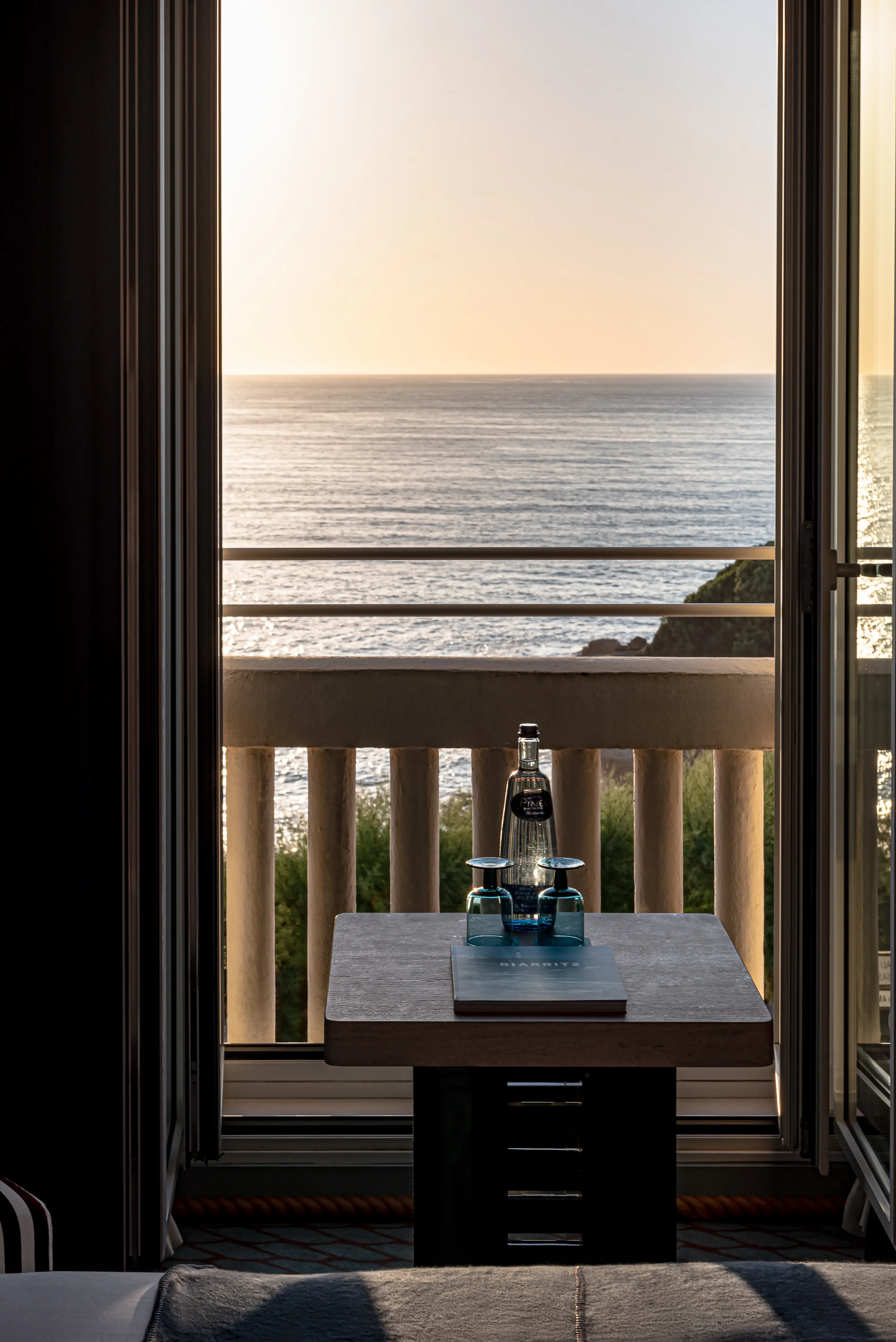 Tavolo con due bicchieri blu e una bottiglia vicino a un balcone aperto con vista sul mare al tramonto.