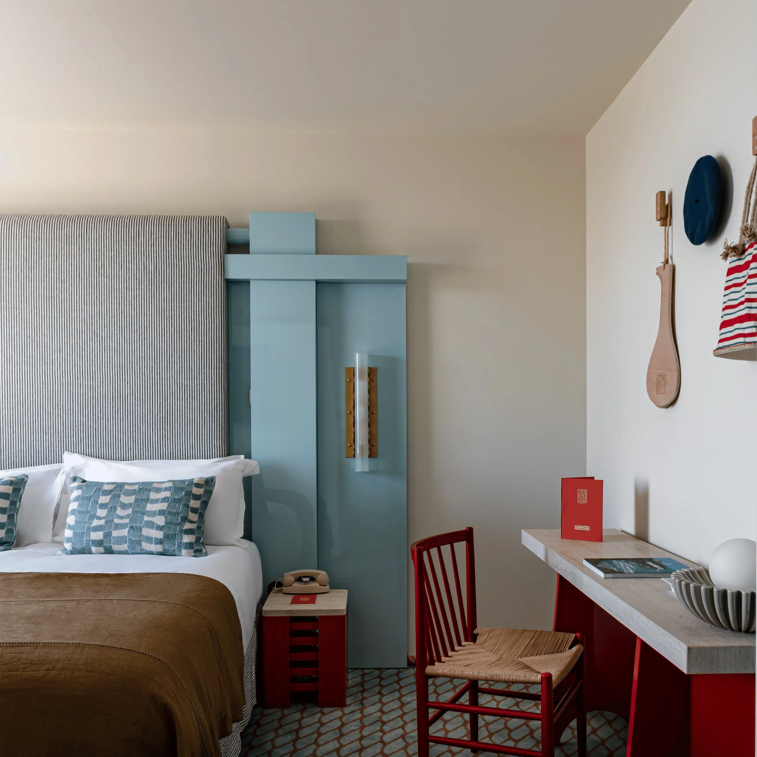 Camera d'albergo con letto, testiera blu, coperta marrone, sedia rossa e scrivania con decorazioni e libri.