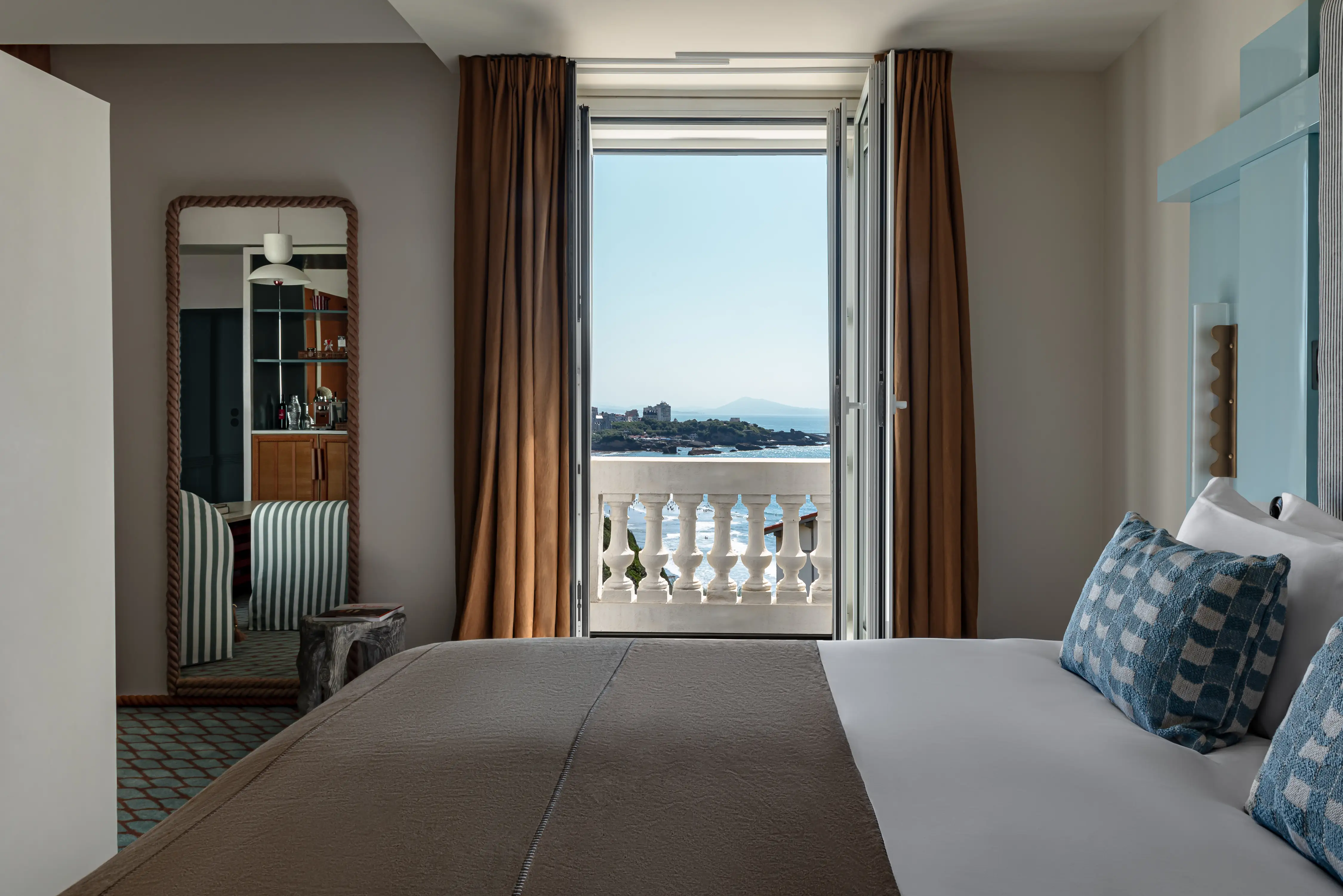 Camera d'albergo con letto, cuscini a righe, grande specchio e porta aperta con vista mare e balcone.