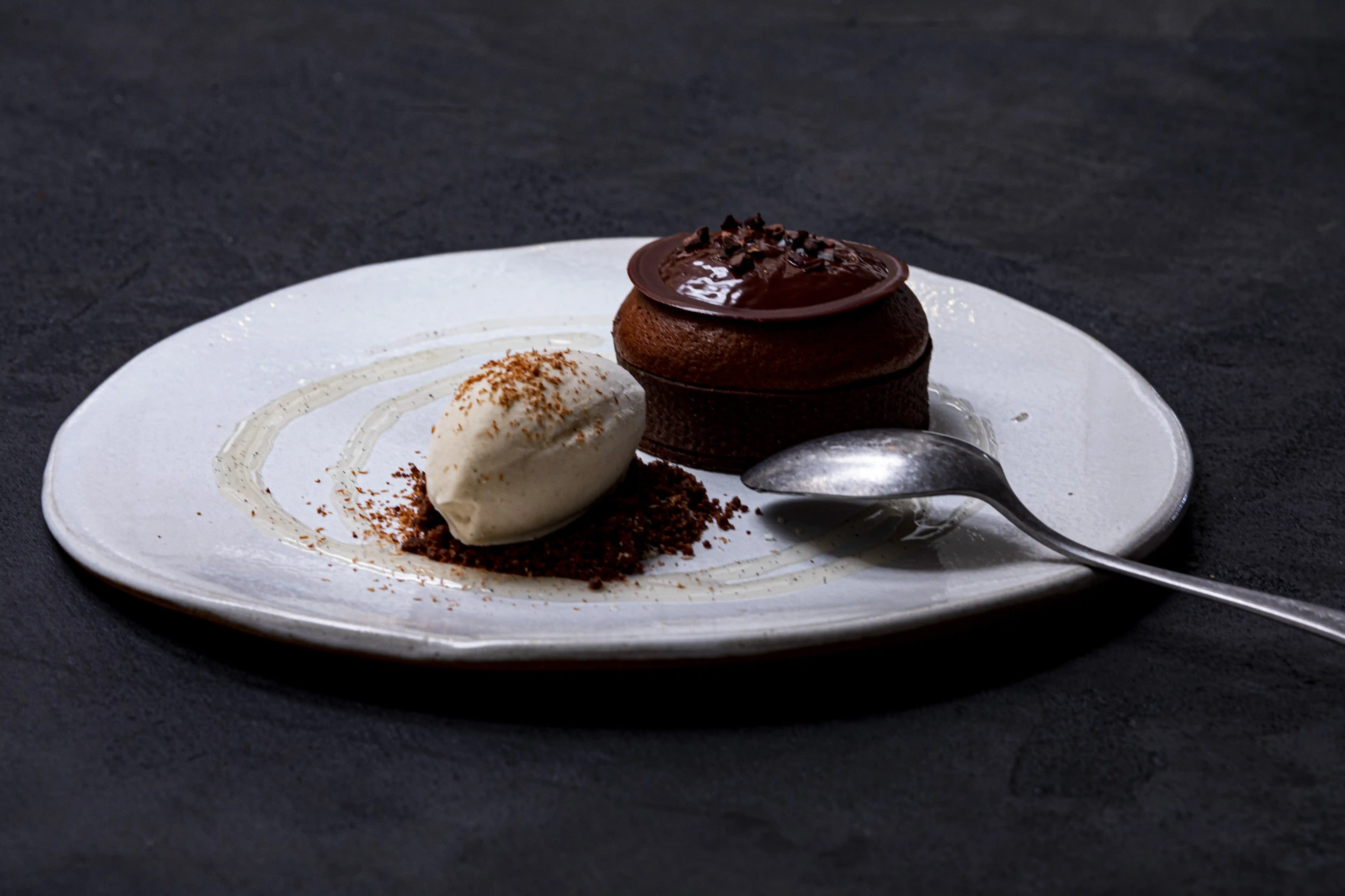 Dessert al cioccolato con una pallina di panna montata e briciole di cioccolato su un piatto bianco con un cucchiaio.