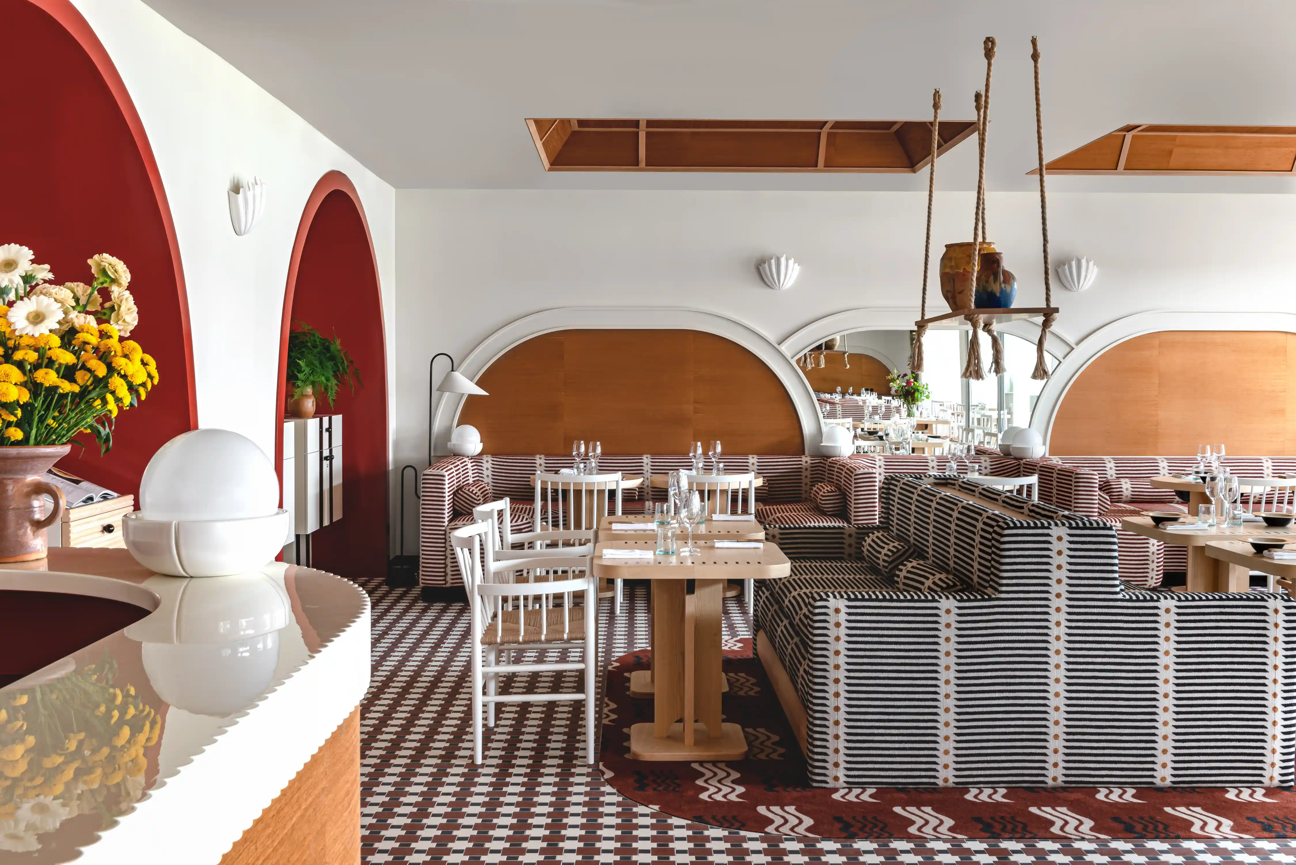 Interno moderno del ristorante con divani a righe, tavoli in legno, sedie bianche e accenti murali ad arco rossi.