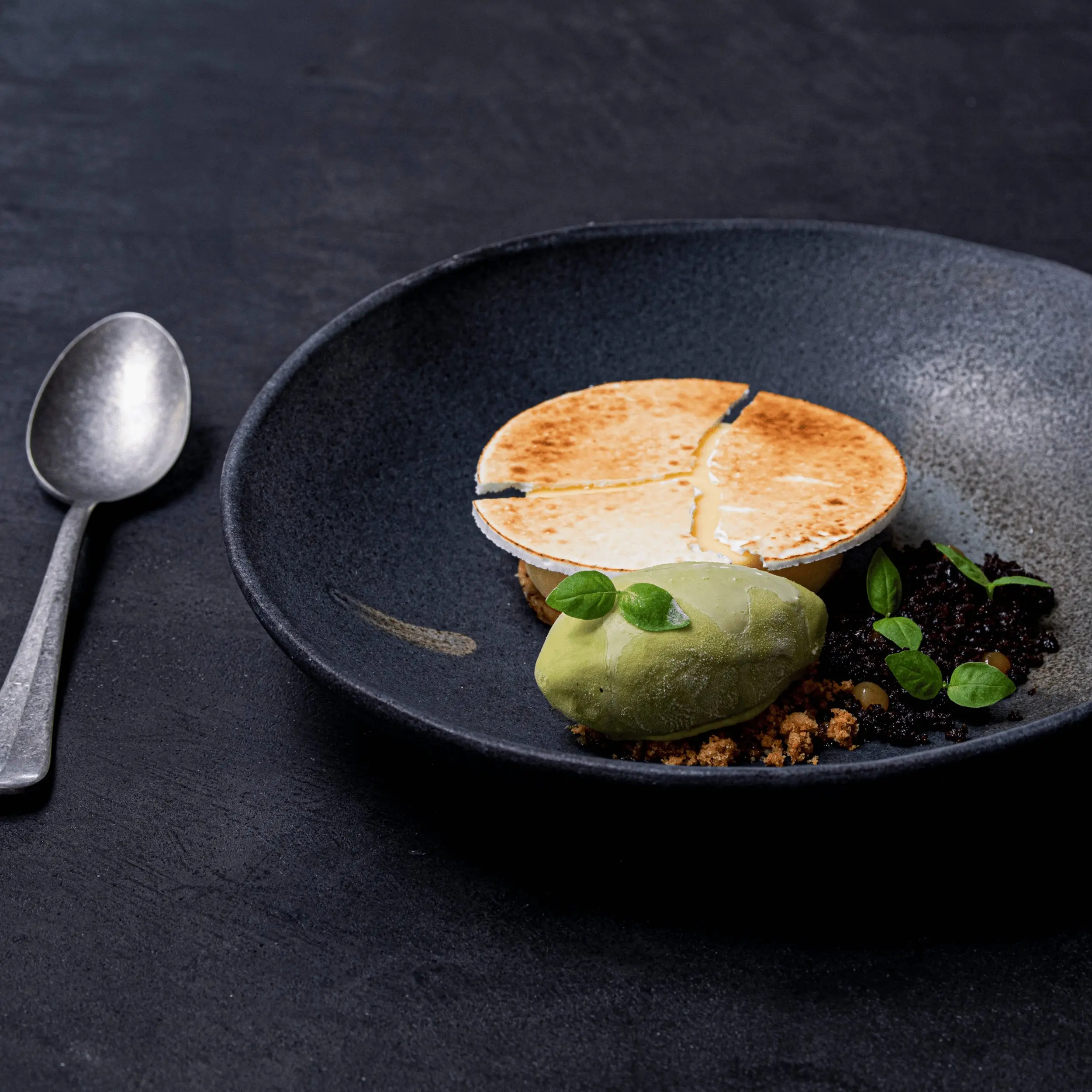 Dessert gourmet con parte superiore tonda tostata, sorbetto verde, briciole ed erbe aromatiche su un piatto nero.