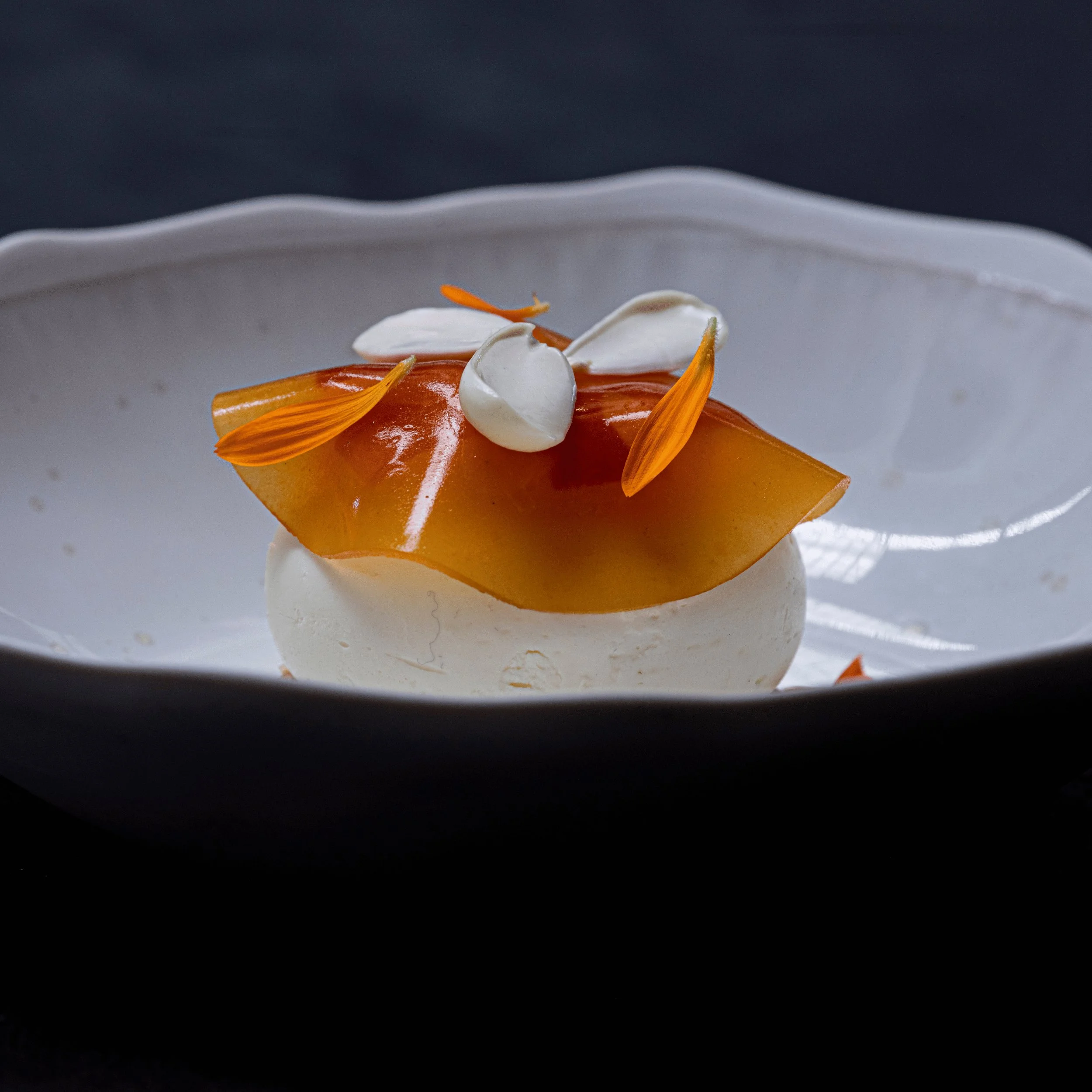 Dessert elegante con sfoglia ambrata traslucida, crema bianca e petali di fiori d'arancio su piatto bianco.