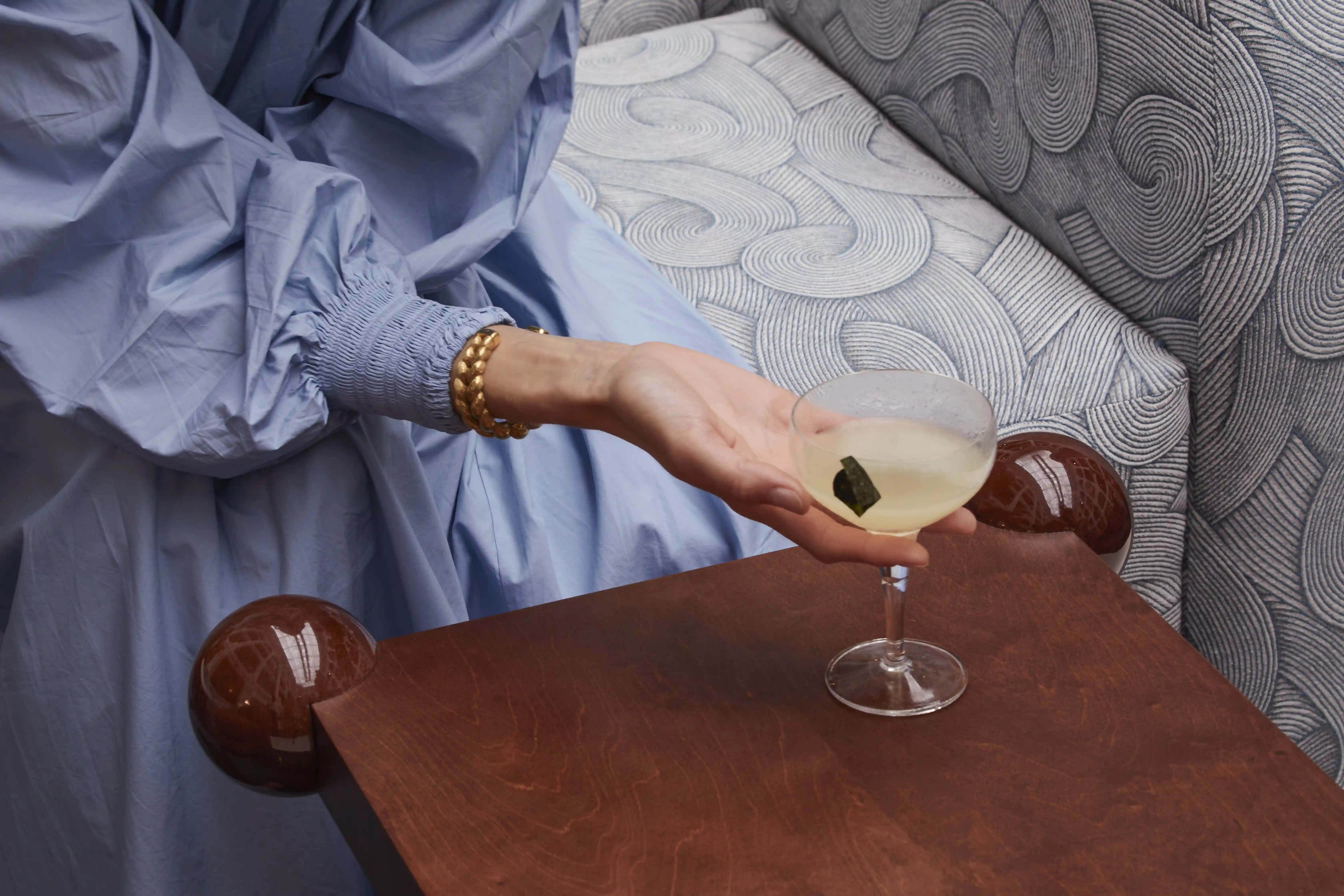 Mano con un grosso bracciale in oro che regge un bicchiere da coupé con un cocktail leggero e una guarnizione.
