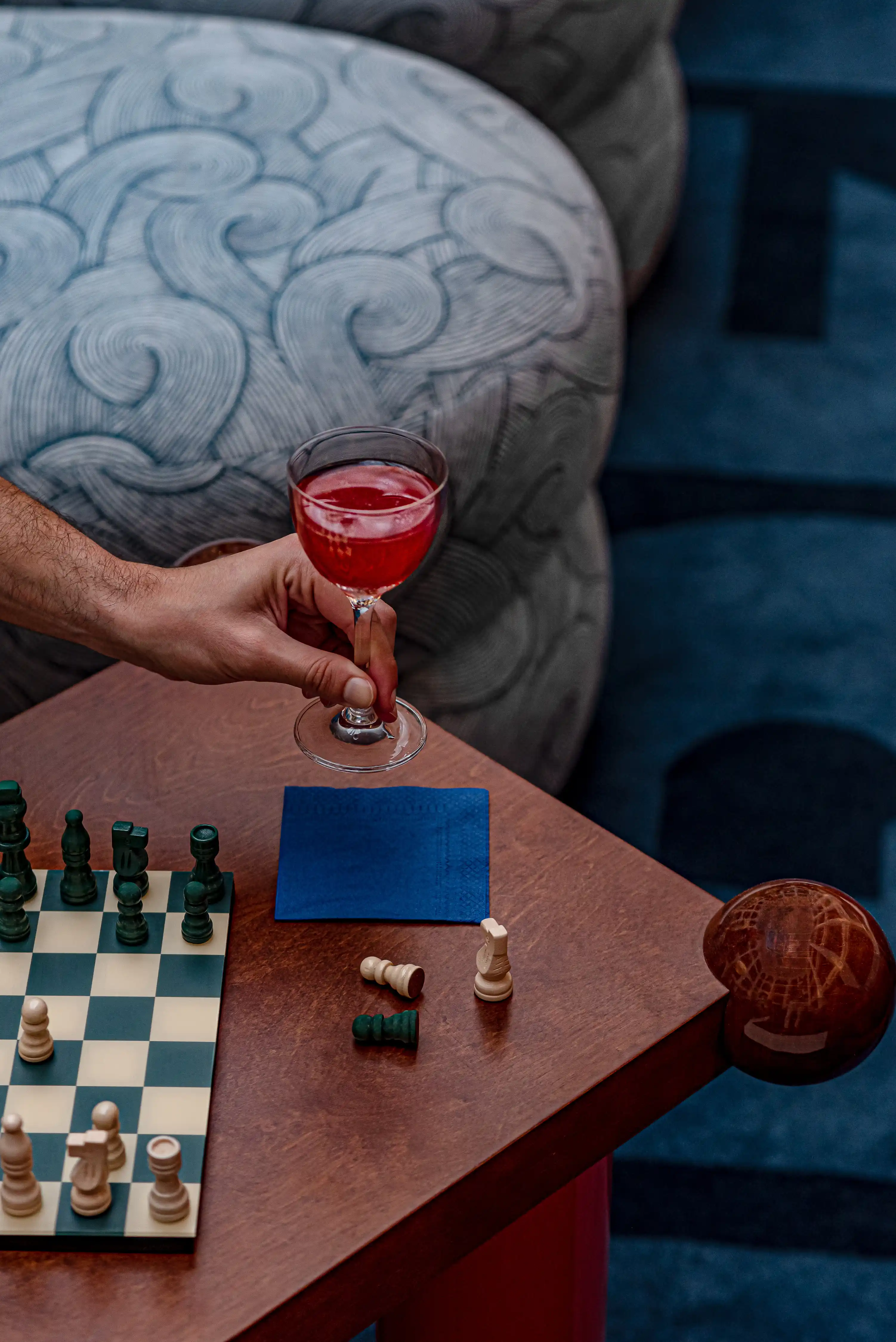 Mano che tiene un bicchiere da cocktail rosso sopra un tavolo con una scacchiera e pezzi degli scacchi caduti.