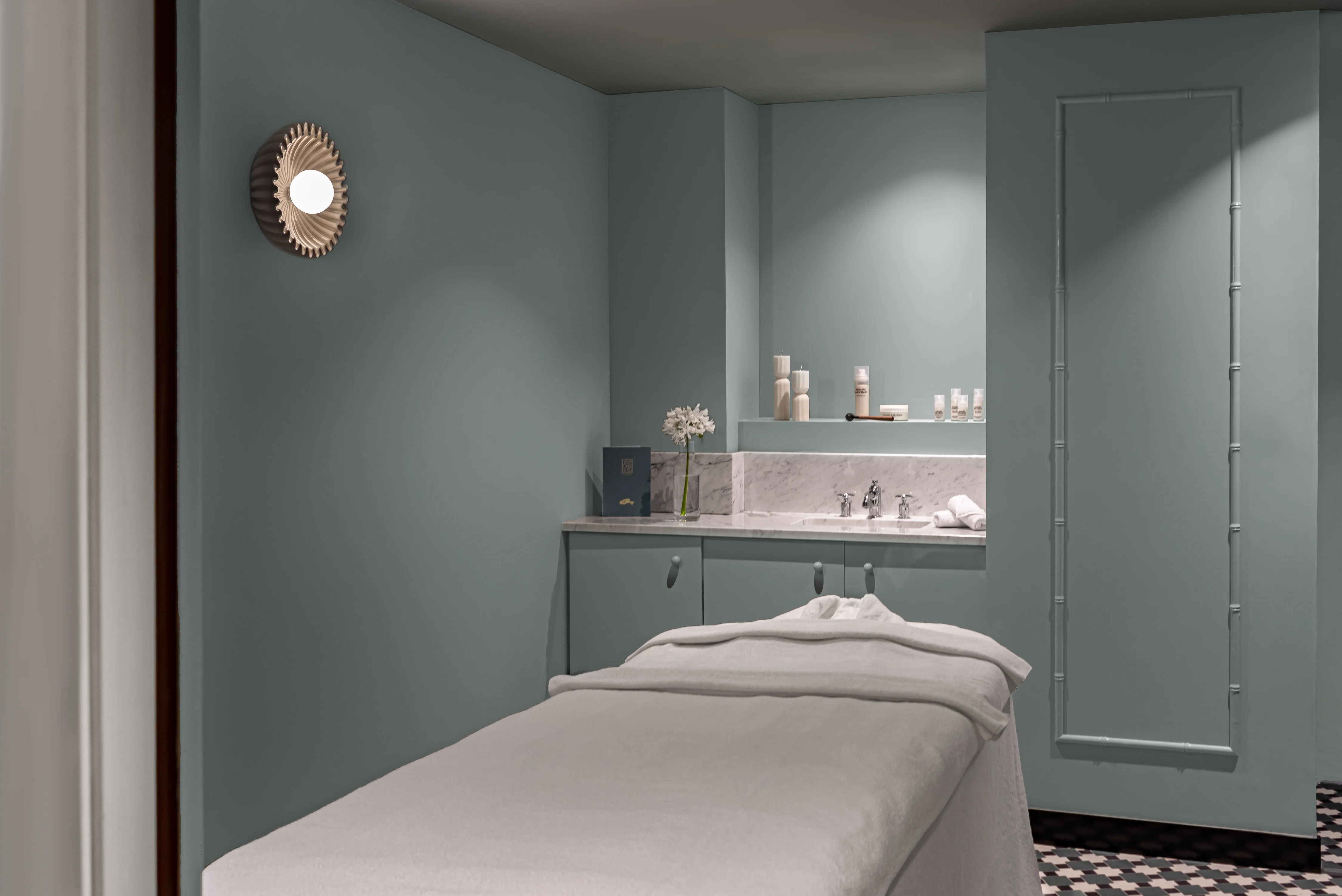 Sala trattamenti spa con lettino da massaggio, morbide pareti grigie, lampada da parete e prodotti per la cura della pelle su bancone in marmo.