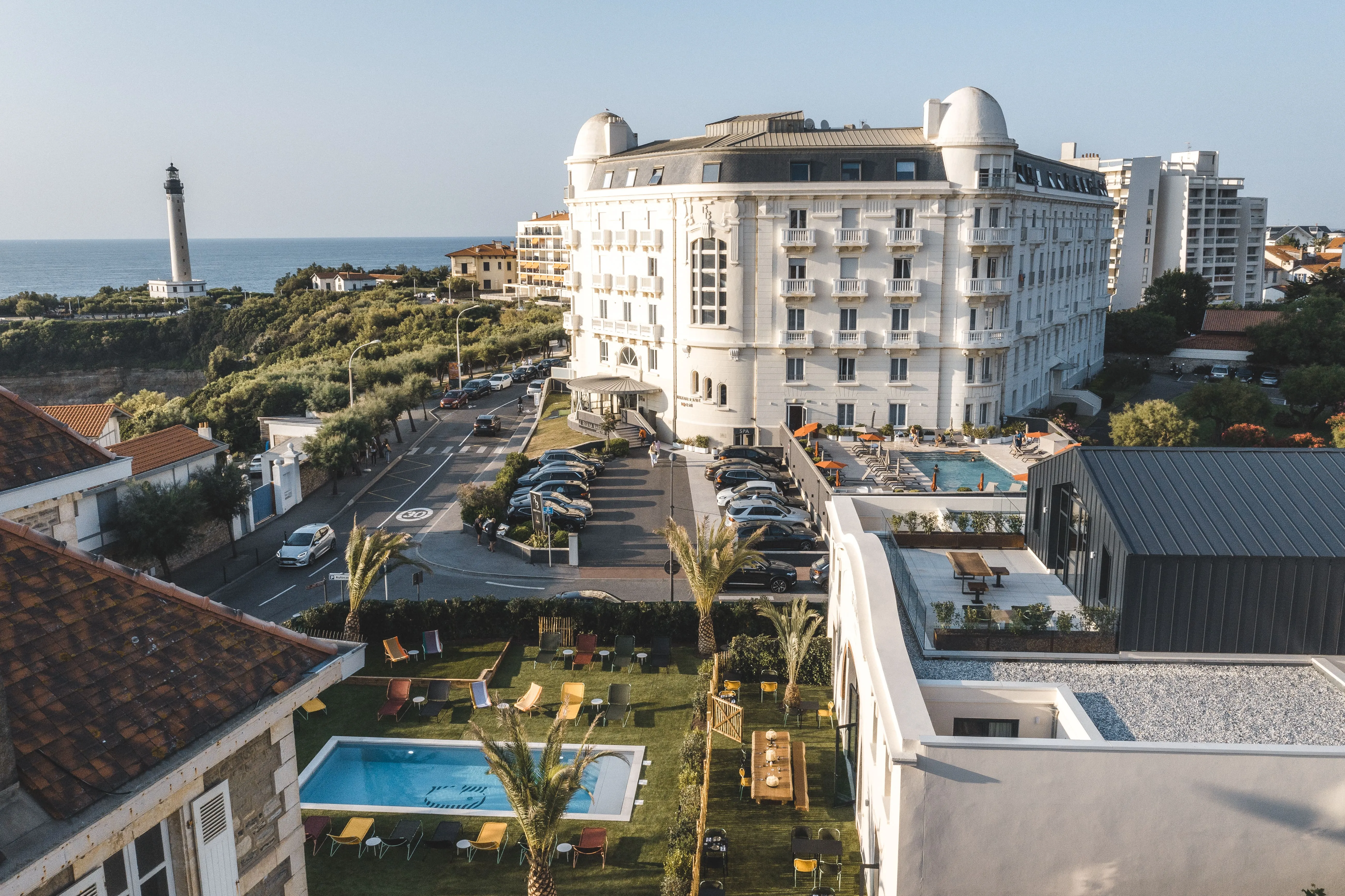 Coastal hotel with pools, palm trees, parked cars, and a lighthouse by the sea in the background.