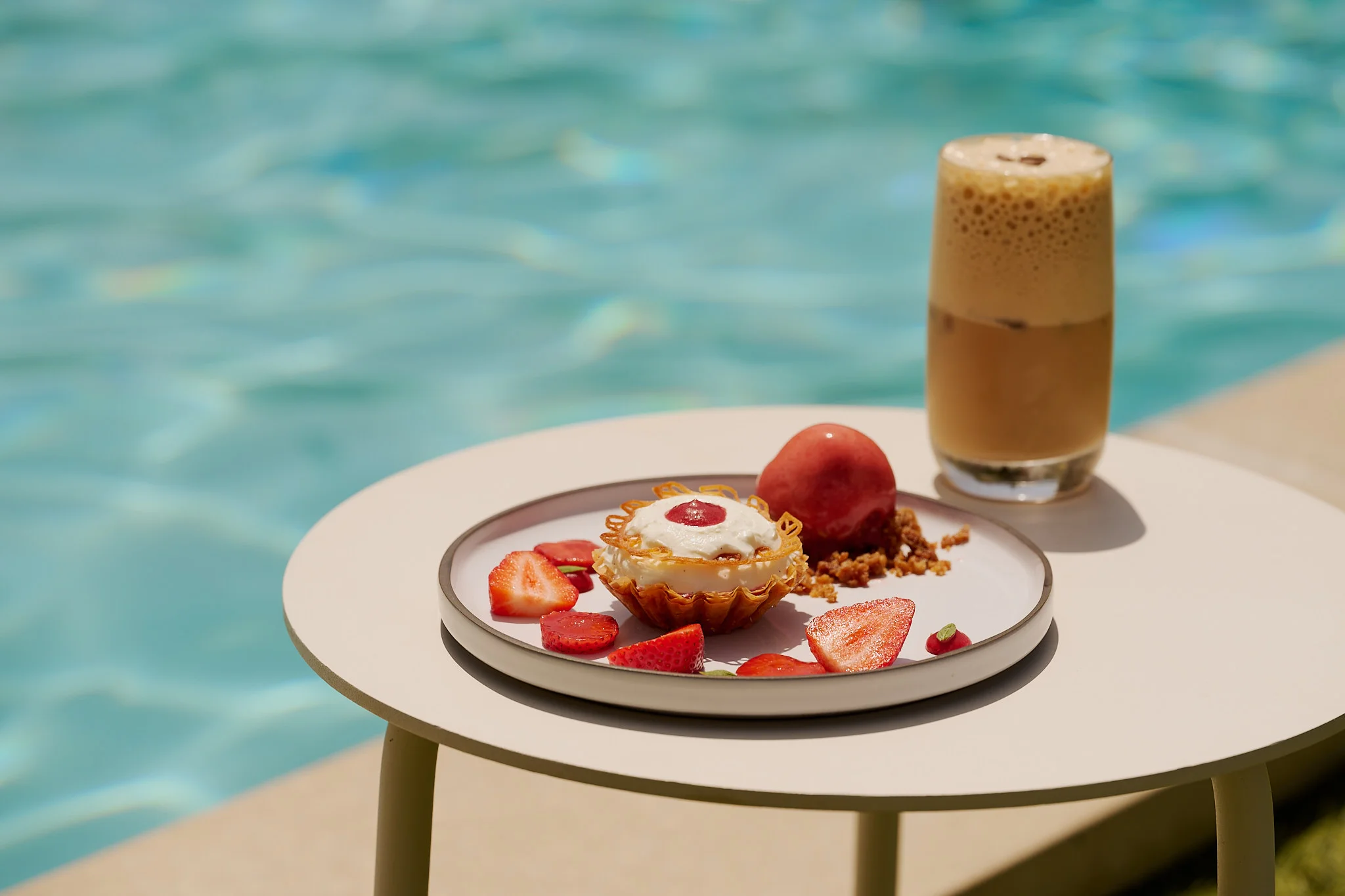 Piatto da dessert con fragole, crostata alla crema, sorbetto alla frutta e caffè freddo a bordo piscina su un tavolo rotondo.
