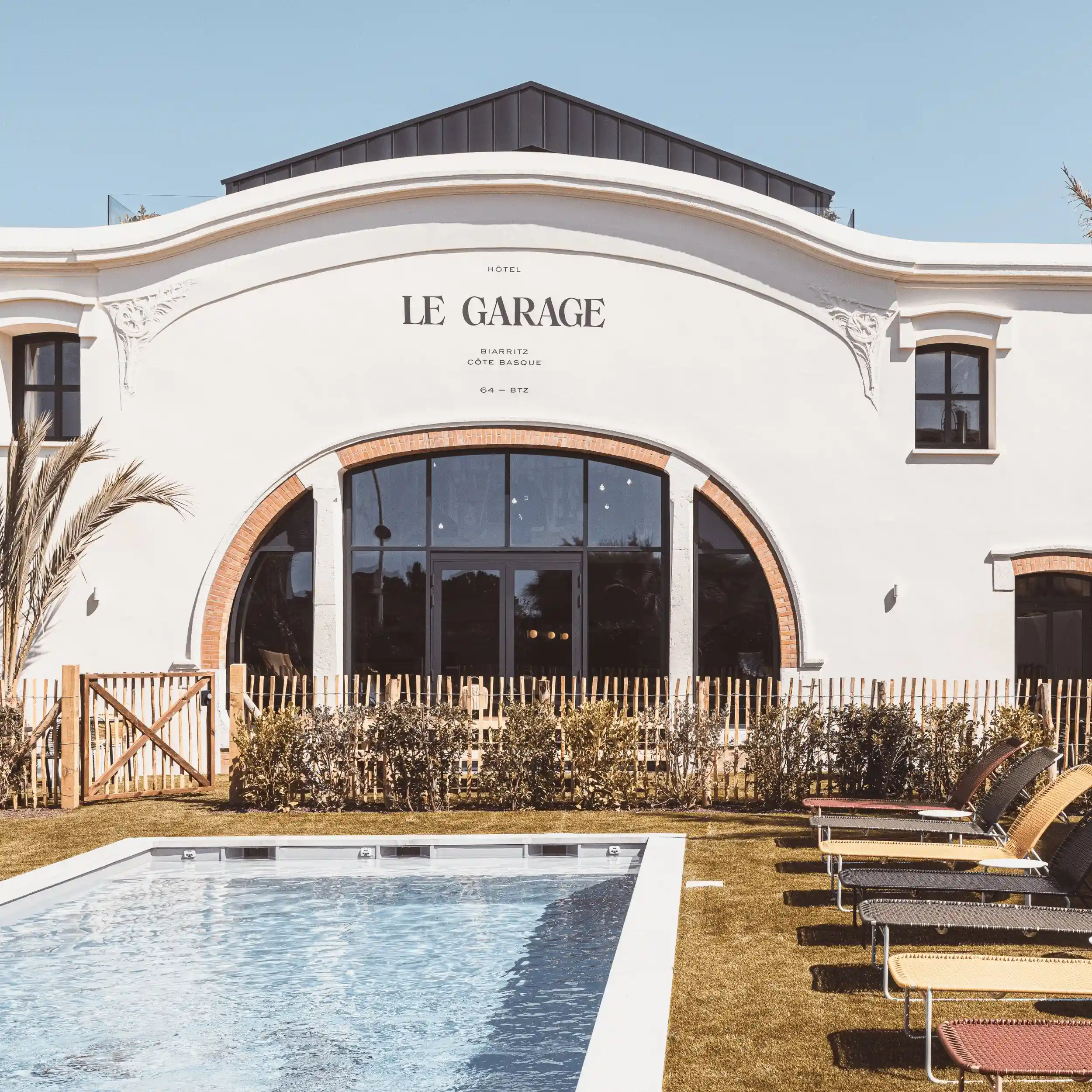 Piscina all'aperto e sedie a sdraio di fronte all'hotel Le Garage con facciata bianca e grande finestra ad arco.