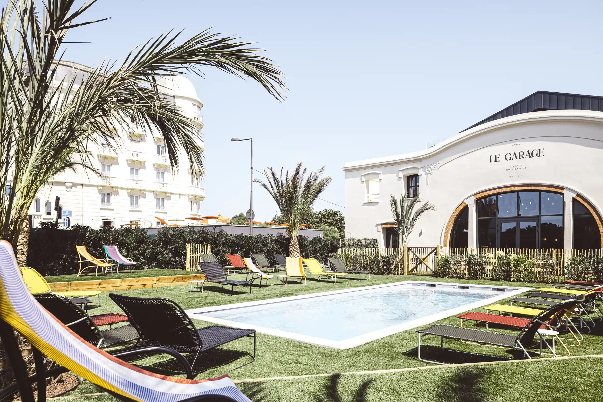 Piscina all'aperto con sedie a sdraio colorate su erba artificiale vicino all'edificio dell'hotel Le Garage e alle palme.