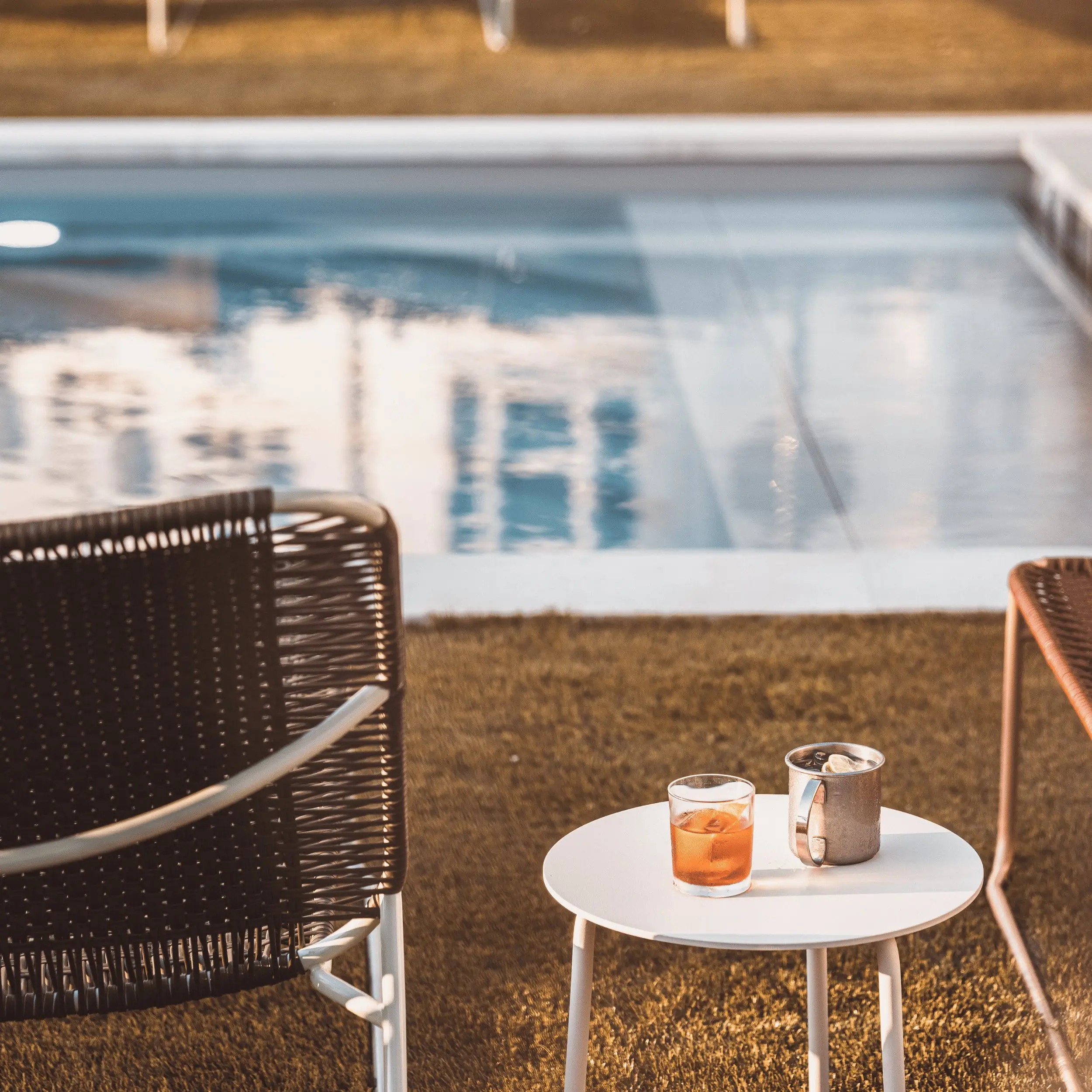 Two chairs and a small white table with two drinks by a pool under warm light.