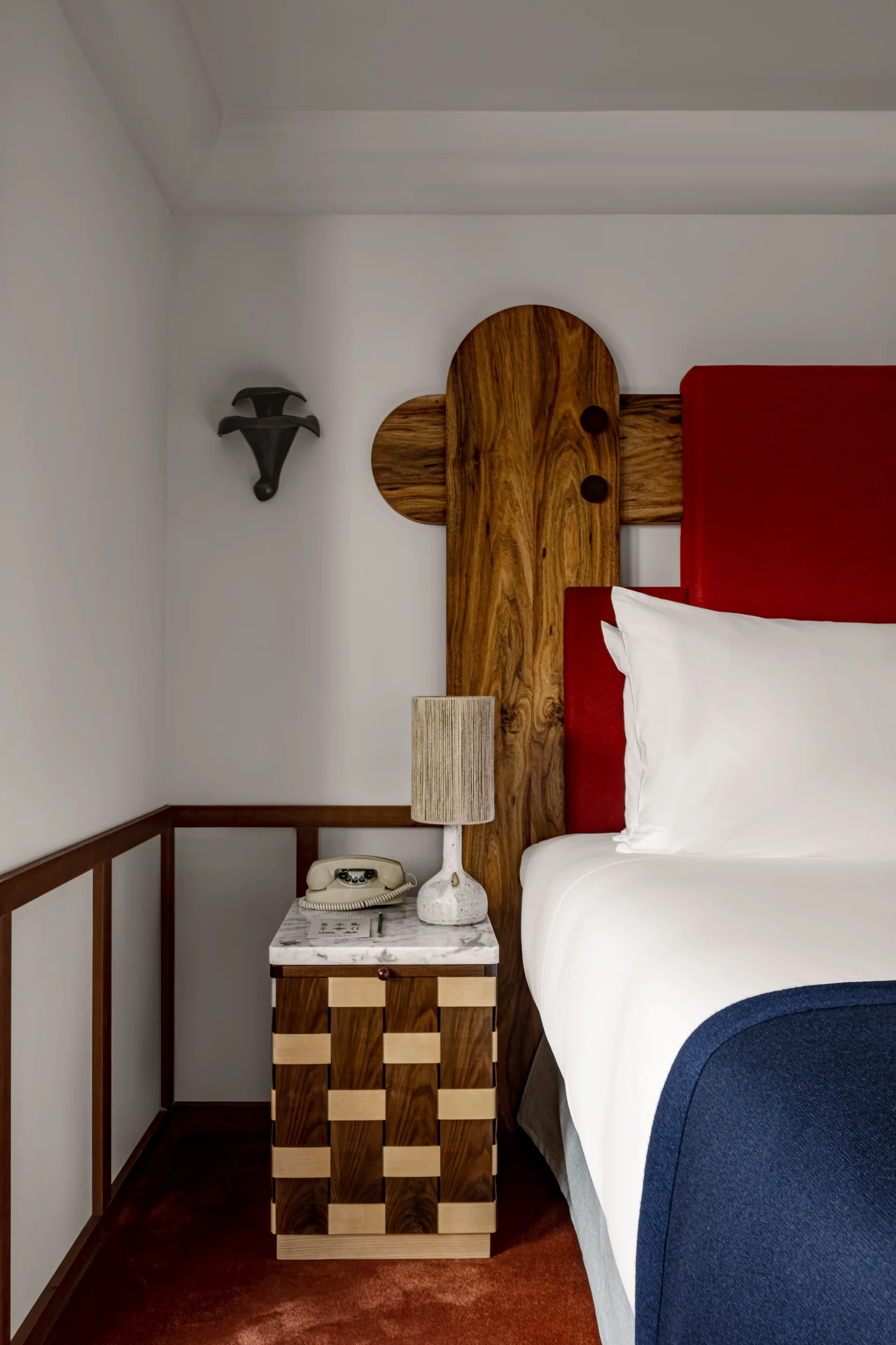 Cama com cabeceira vermelha ao lado de uma mesa de cabeceira de madeira segurando uma lâmpada e um telefone rotativo antigo.