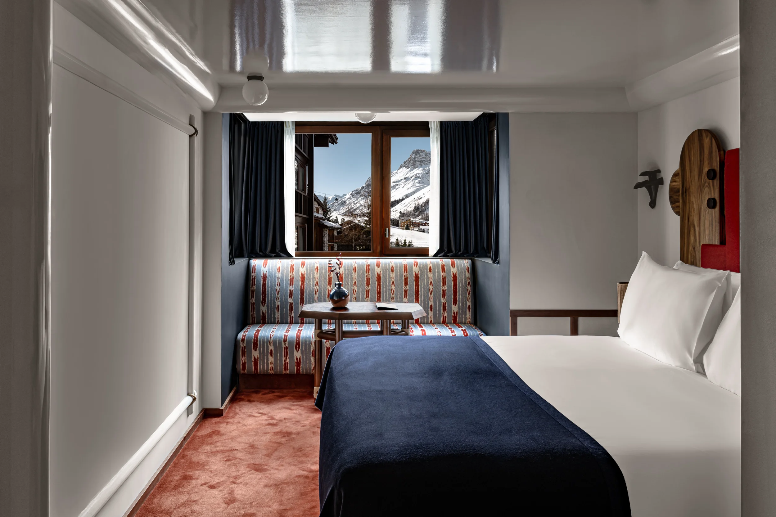 Quarto de hotel com cama, banco estampado, mesa pequena e janela com vista para a montanha nevada.